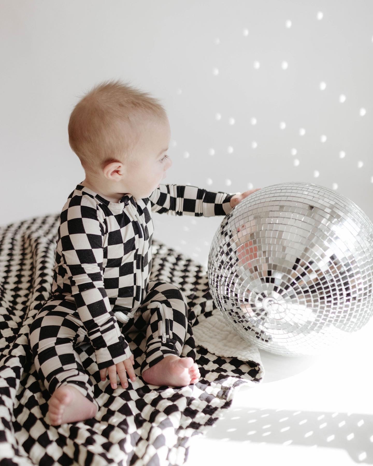 A baby dressed in Forever French Baby’s Bamboo Zip Pajamas in Black Checkerboard, featuring convenient rollover cuffs, sits on a coordinating checkered blanket while touching a large disco ball. The light reflections from the disco ball create dots of light on the white wall in the background, perfectly capturing the essence of Forever French Baby.