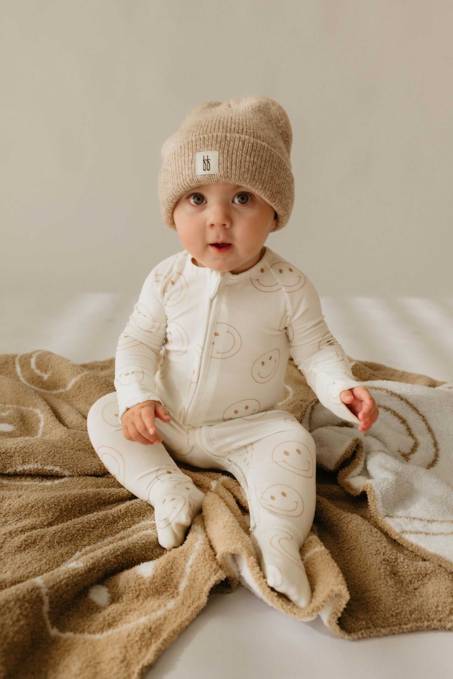 A baby in a beige knit beanie and white onesie with smiley faces from forever french baby sits on the Plush Blanket | Original Just Smile by forever french baby, looking at the camera against a neutral background.