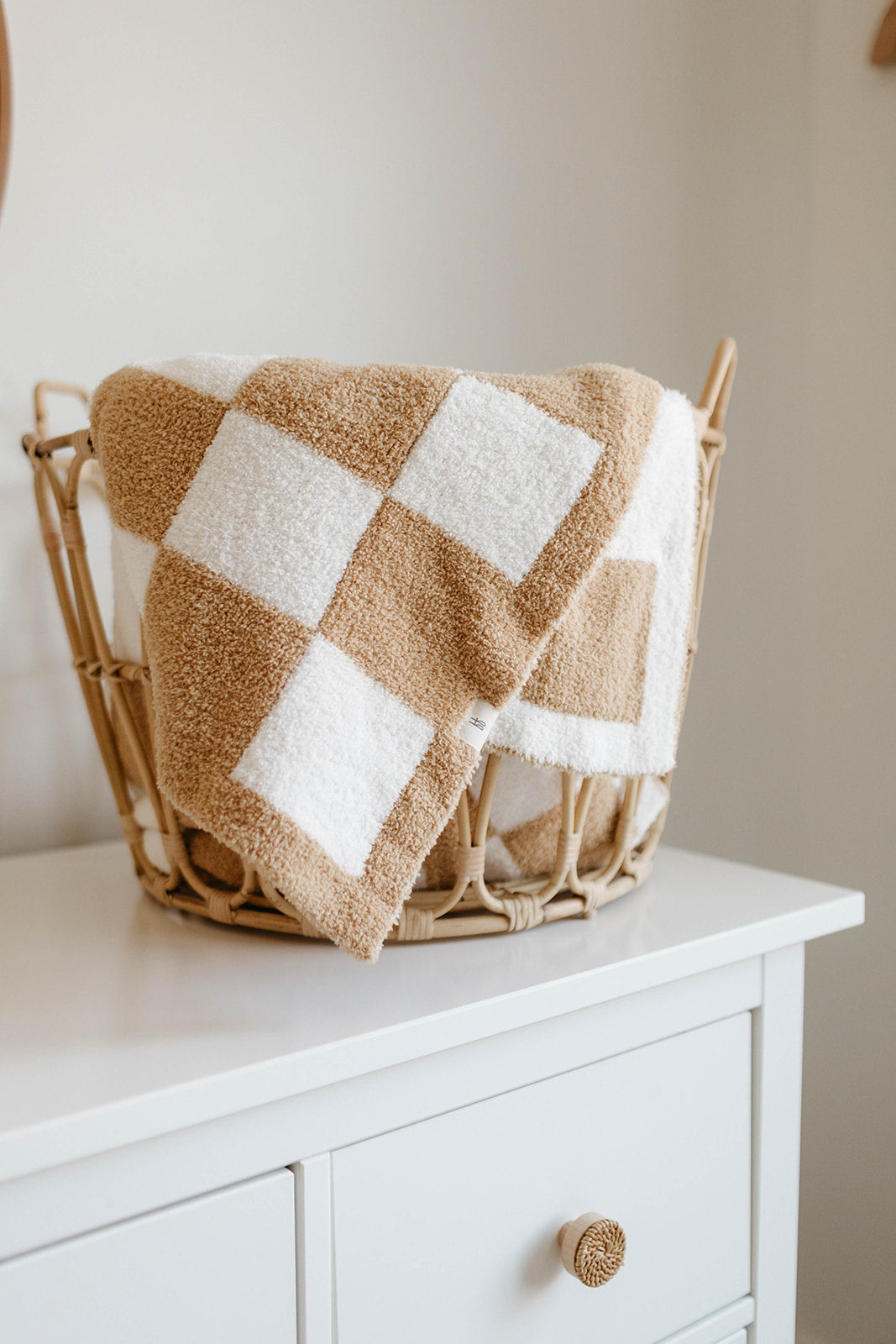 A Plush Blanket | Original Checkerboard by forever french baby, made of OEKO Standard 100 microfiber feather yarn, is draped over the edge of a wicker basket on top of a white dresser. The beige and white checkered pattern, combined with its cozy texture, creates a warm, minimalist feel in the room.