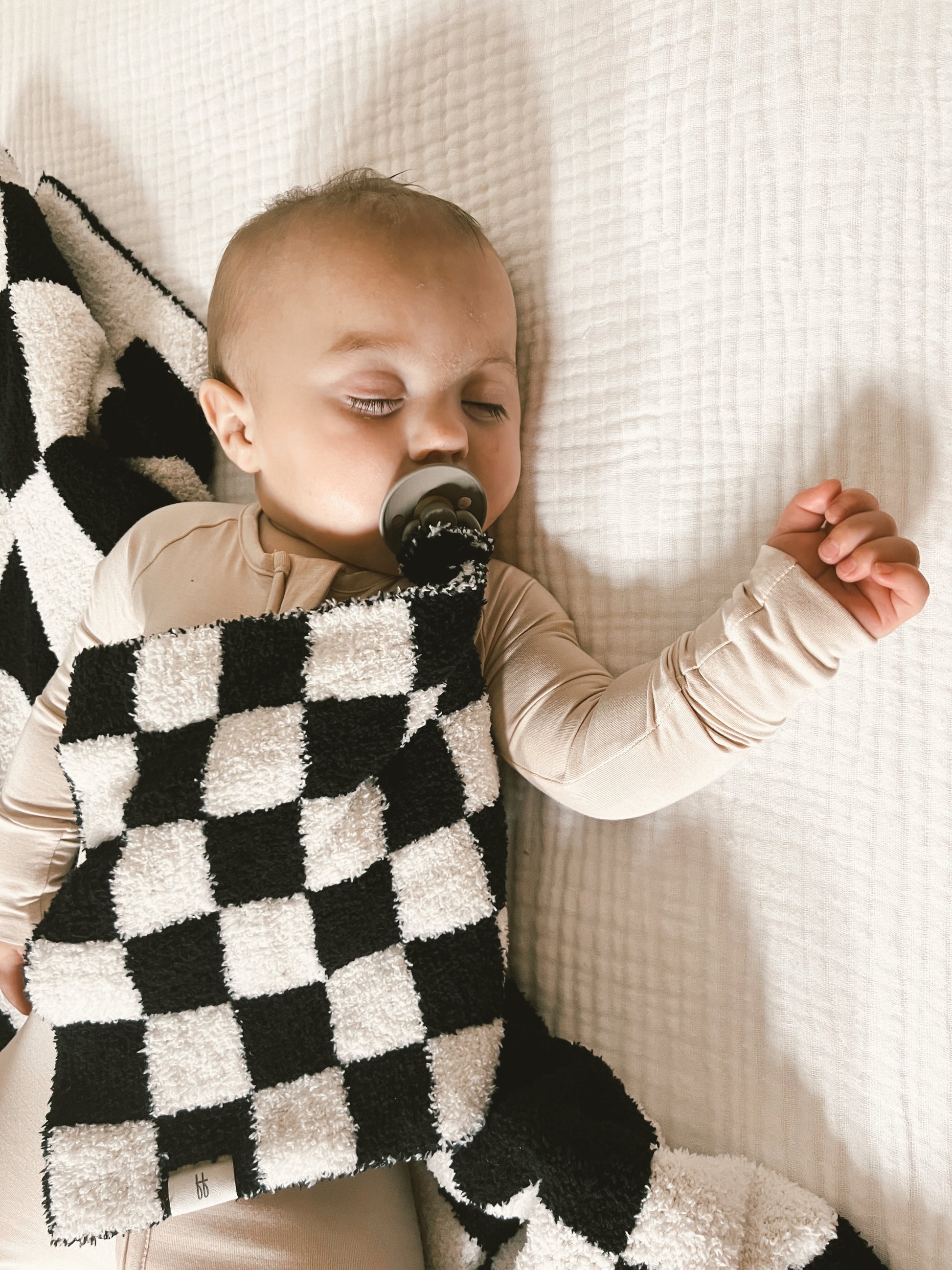 A baby sleeps peacefully with a black pacifier in their mouth, lying on a white bed. They are swaddled in a Lovey | Black & White Checkerboard blanket from forever french baby, with one arm protruding outside, resting on the blanket. The baby's outfit matches the light, neutral tones of the blanket.