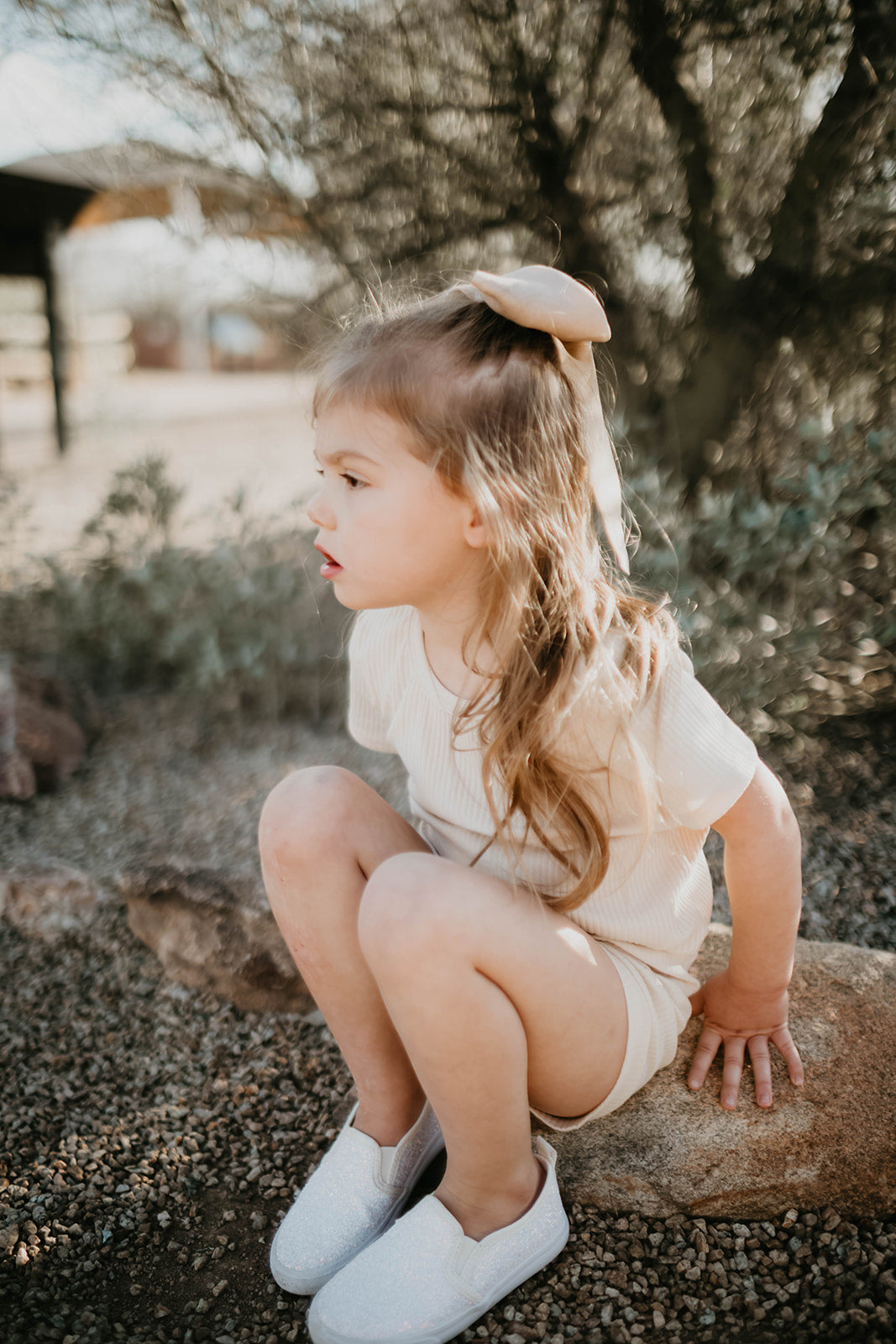 A young girl with long brown hair and a bow sits on a rock outdoors, wearing the Ribbed Set - Vanilla from forever french baby. Her white shoes complement the light-colored ribbed cotton outfit against a backdrop of blurred greenery and a wooden structure.
