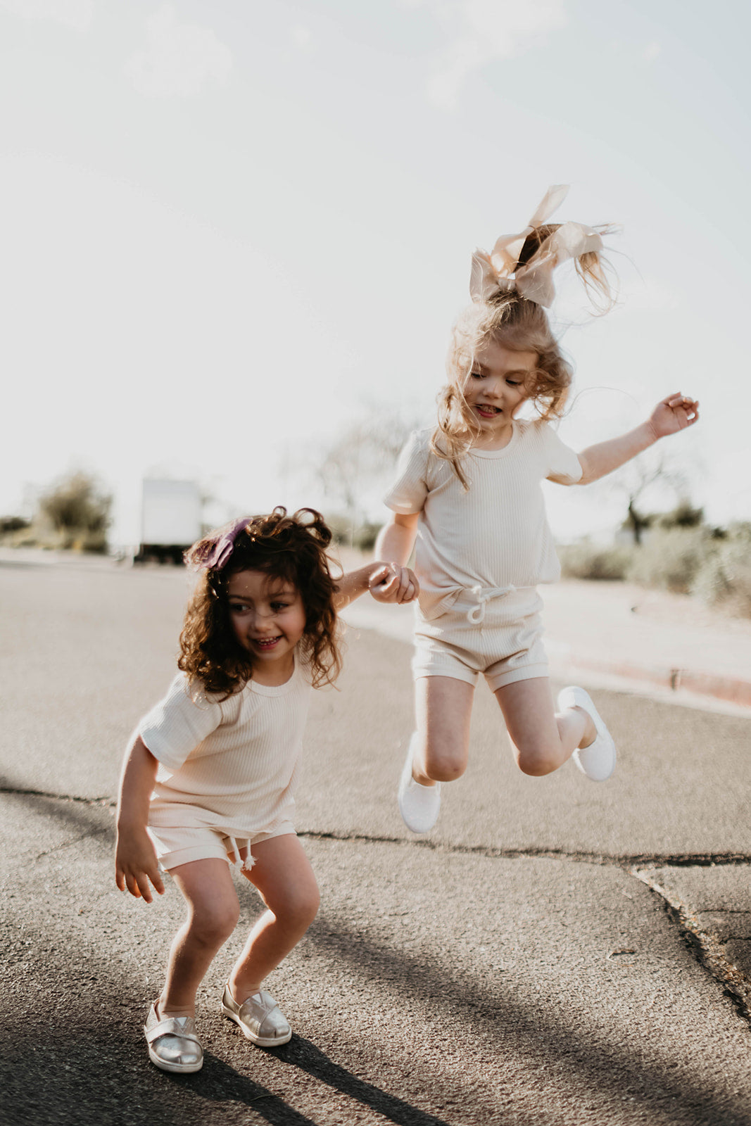 Two young children joyfully play on a sunny street in the Ribbed Set - Vanilla by forever french baby. One jumps with a big bow in their curly hair while holding the other's hand, both smiling against blurred trees and sky.