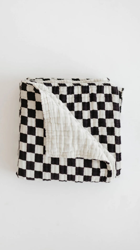 A folded Muslin Quilt | Black & White Checkerboard from forever french baby with one corner flipped to reveal a plain white backside sits on a white background. Made from soft muslin cotton, the quilt is neatly arranged, showcasing its contrasting patterns and textures, perfect for adding charm to your Forever French Baby collection.