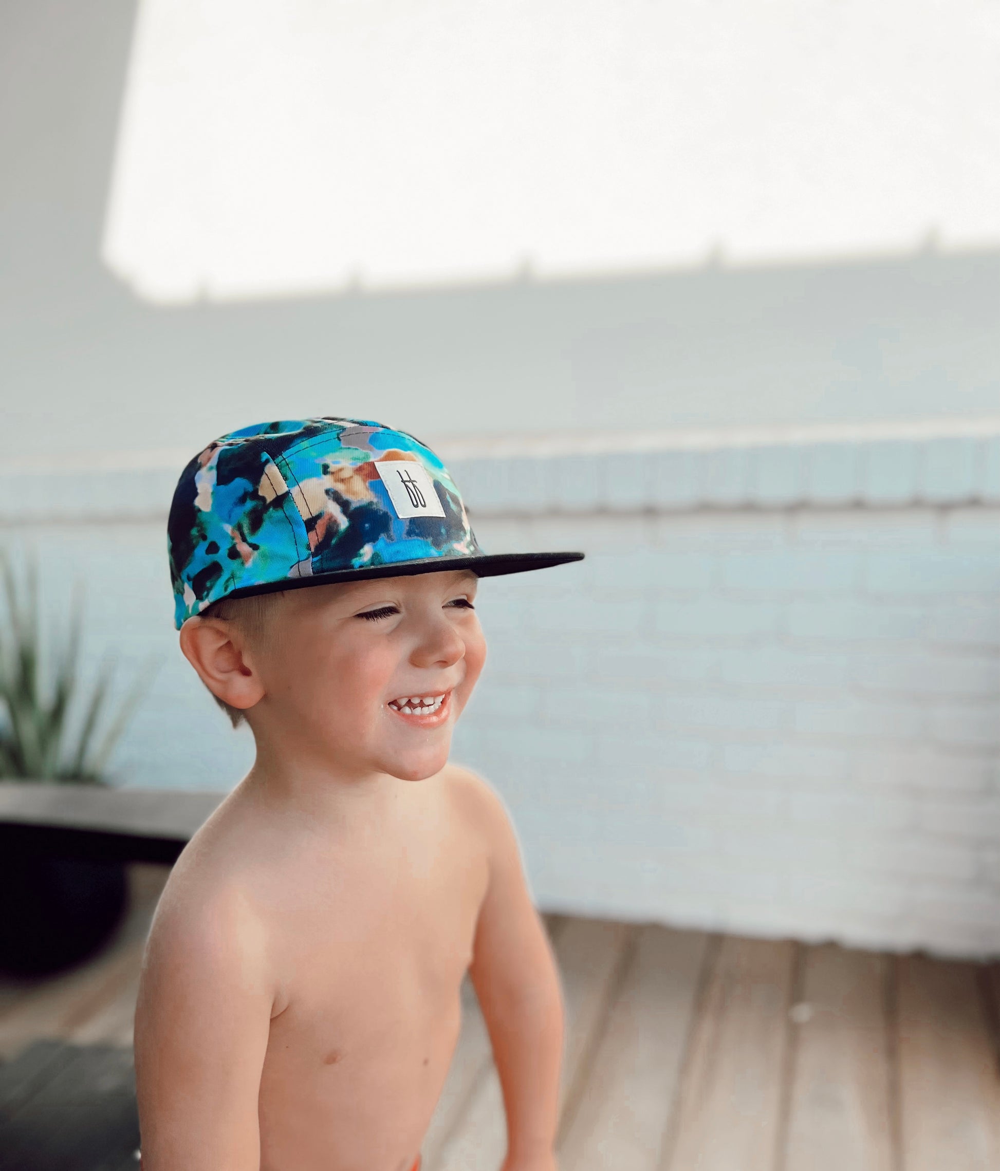 A young child stands shirtless, smiling brightly, and wearing the Snap Back | Charli Print cap by forever french baby. The background features a white brick wall and a potted plant. Sunlight casts soft shadows on the scene, giving it a warm and cheerful atmosphere.