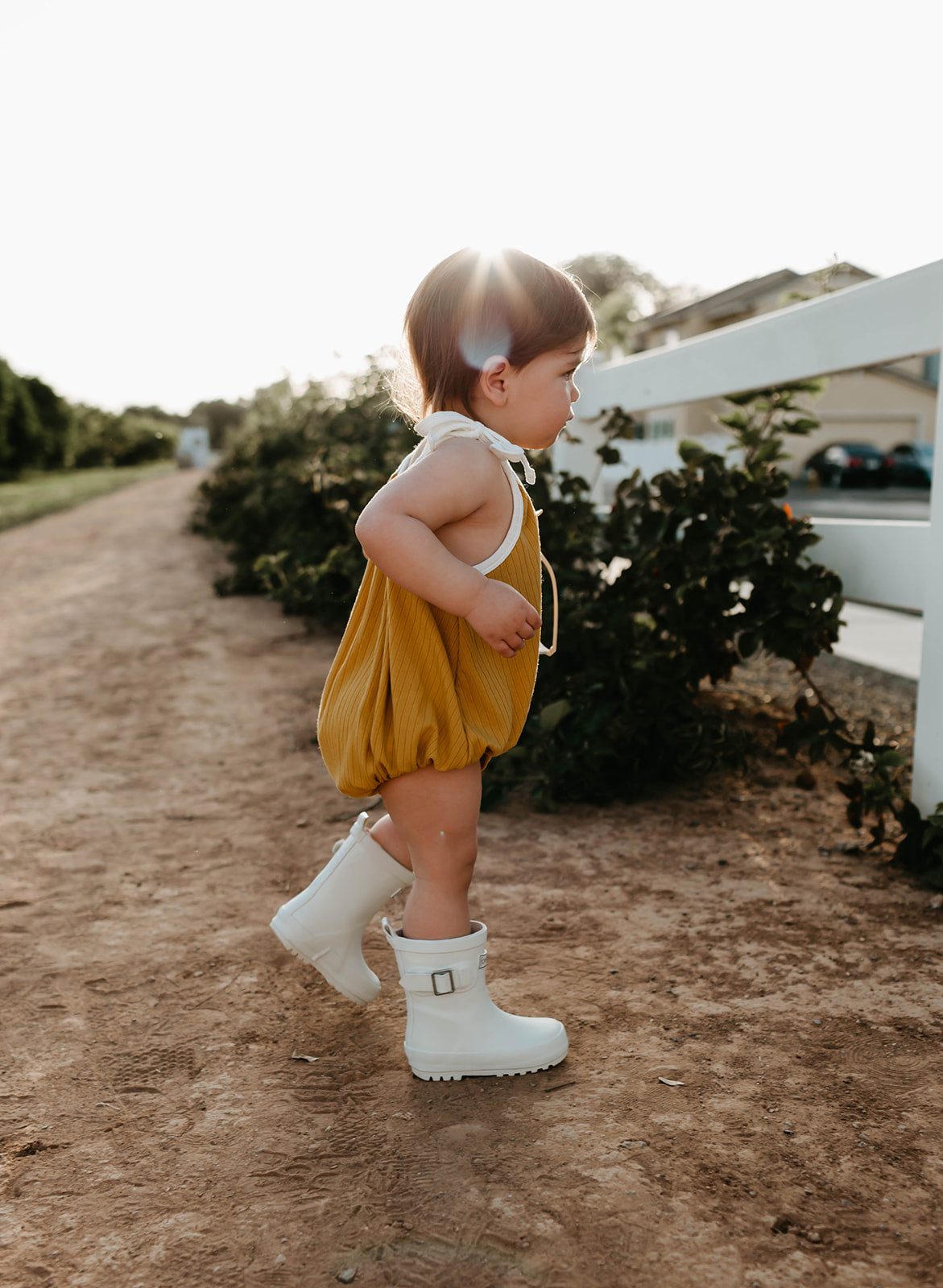 A small child with short brown hair, wearing a yellow romper and the ff Rain Boots in cream by forever french baby, stands on a dirt path next to a white fence and green bushes. The sunlight creates a halo effect around the child's head, highlighting the charm of their all-weather kids footwear.