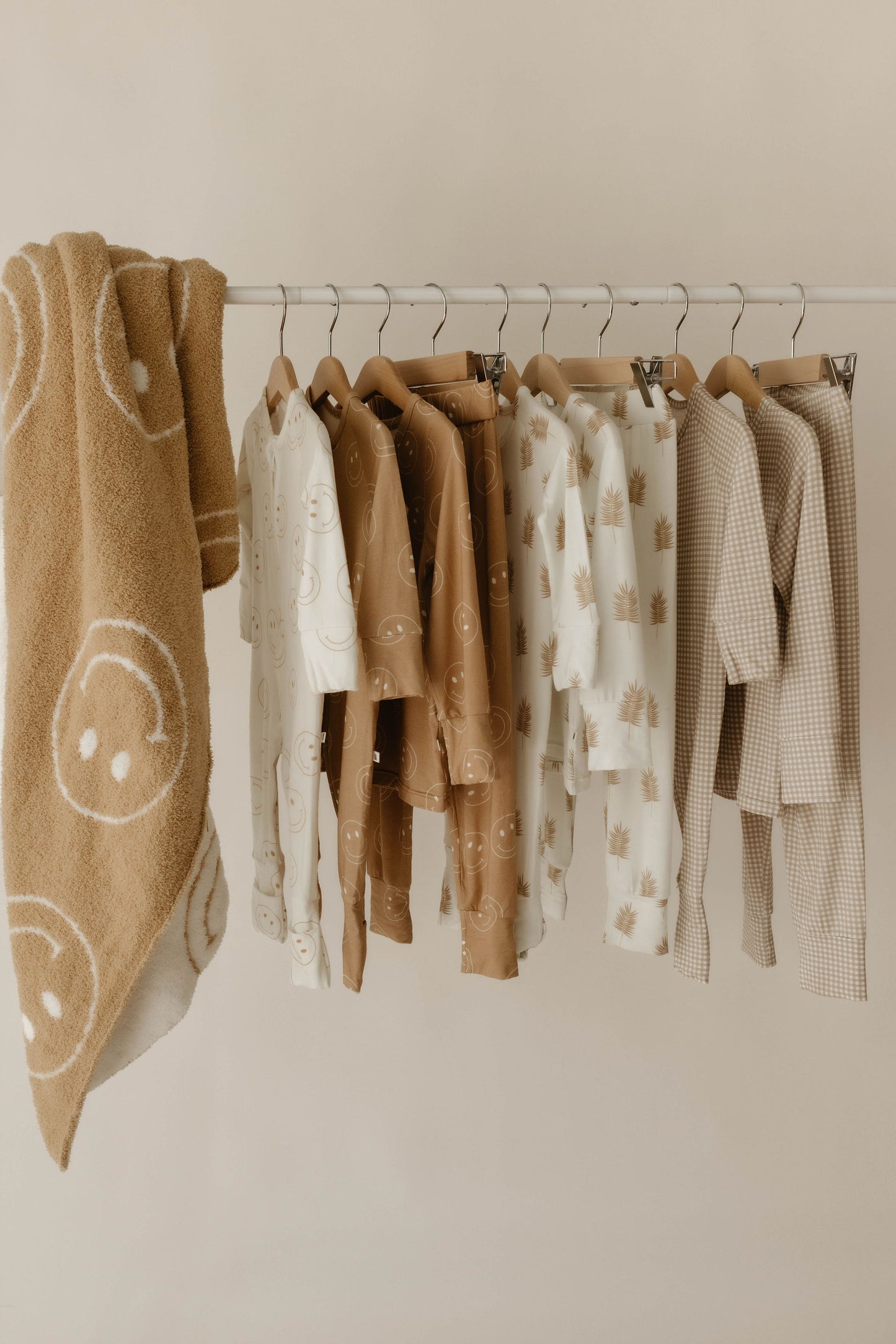 A clothing rack holds neutral-toned children’s clothes with smiley faces, palm trees, and checks next to the forever french baby Plush Blanket | Original Just Smile—dreamy soft with big white smiley faces—set against a plain light beige wall.
