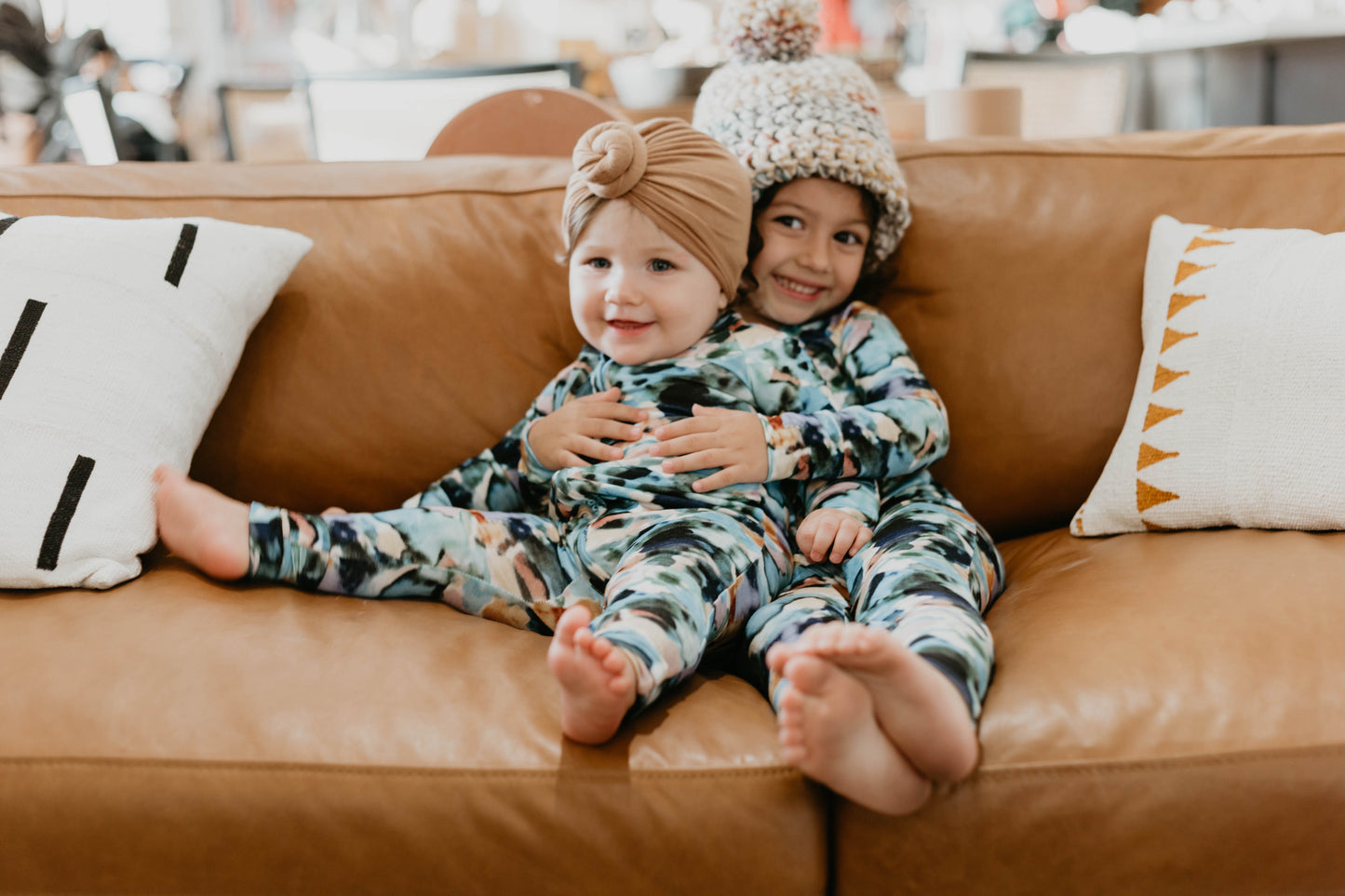Two small children wearing matching Charli Print Bamboo Zip Pajamas from forever french baby sit on a brown leather couch. The older child, donning a knit hat, embraces the younger sibling, who has a head wrap. They are smiling and seem to be reveling in their time together, radiating pure joy in their cozy bamboo clothing.