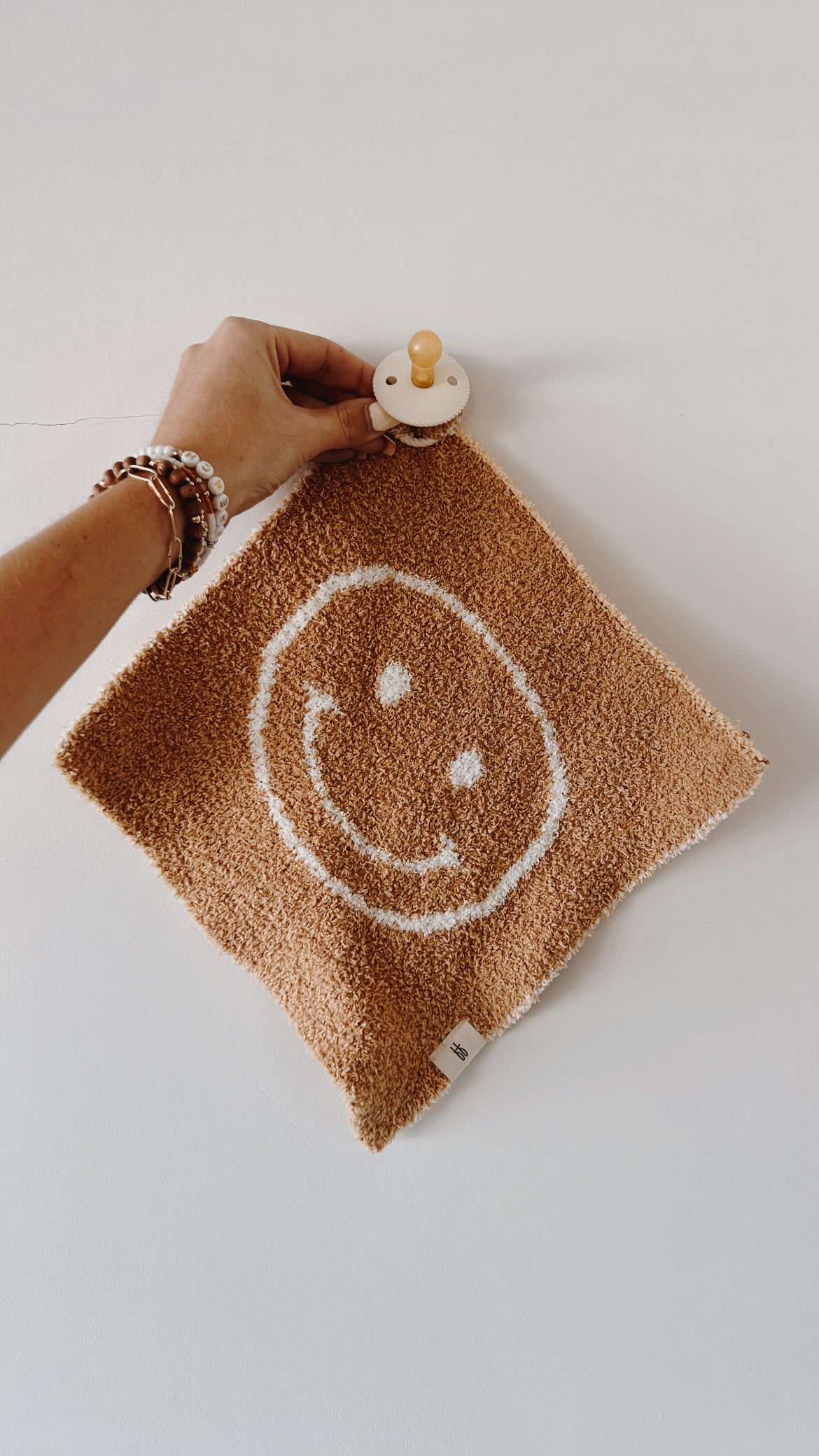 A hand, adorned with beads and a string bracelet, holds the Lovey | Just Smile Tan from forever french baby. This small square brown towel features a white smiley face design and is made of soft microfiber feather yarn. It hangs on a wooden knob attached to a plain light-colored wall, showcasing its cozy charm.
