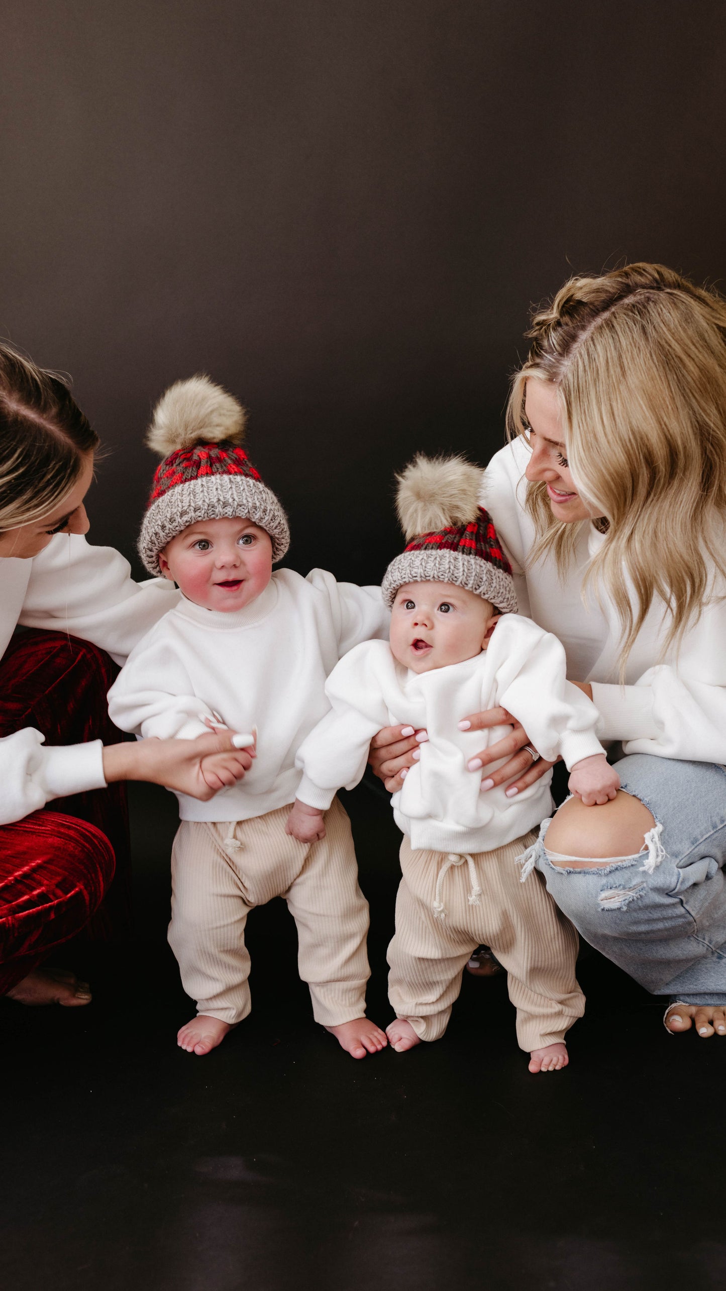 Two women kneel beside two smiling babies in front of a dark background. The babies wear matching forever french baby Ribbed Joggers with elastic waistbands, white sweaters, and red plaid hats with furry pom-poms.