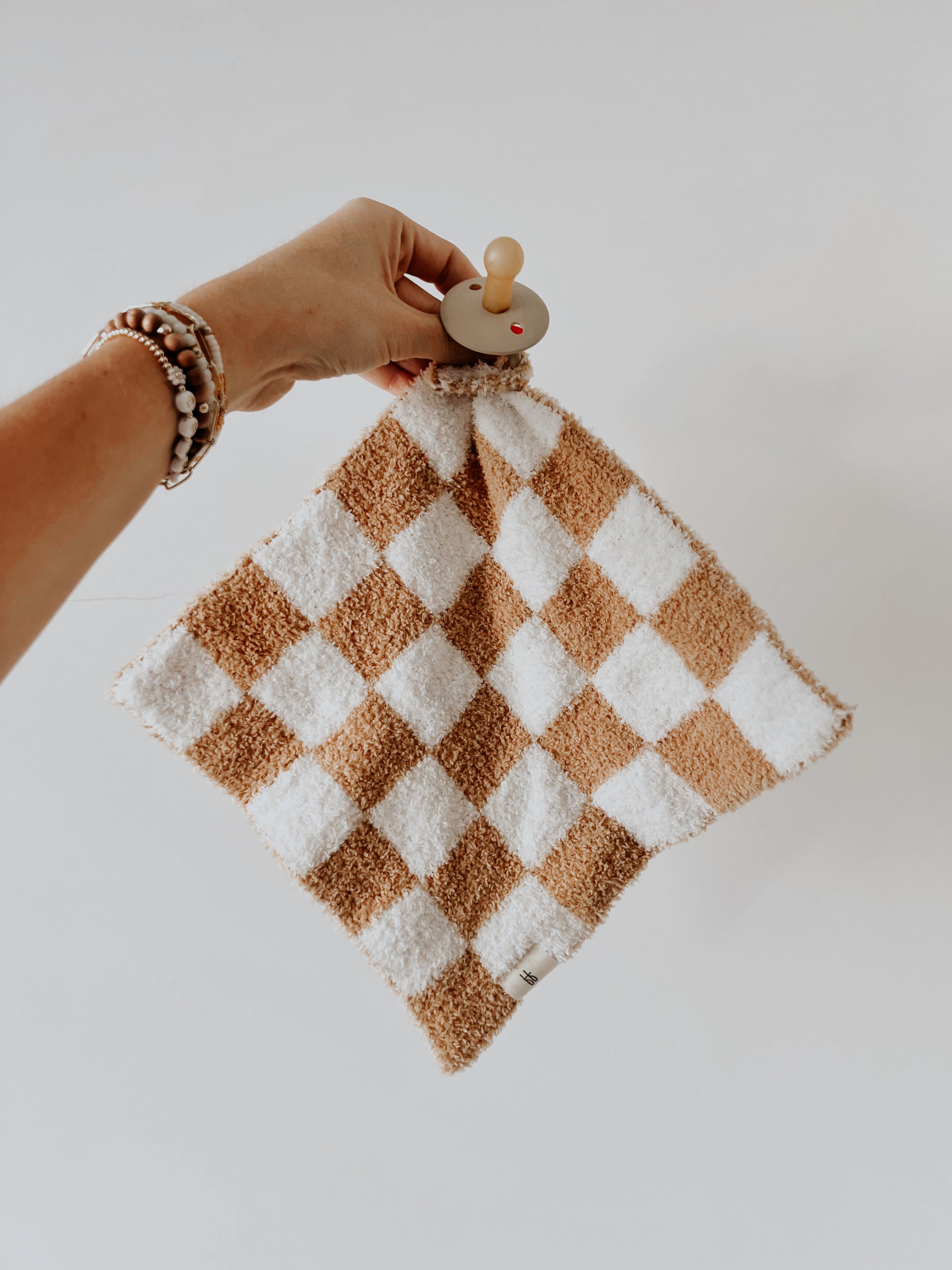 A hand holding a peg with the Lovey | Original Checkerboard from forever french baby, featuring alternately colored squares in white and brown. The person, adorned with multiple beaded bracelets, gently holds the accessory against a plain white background.