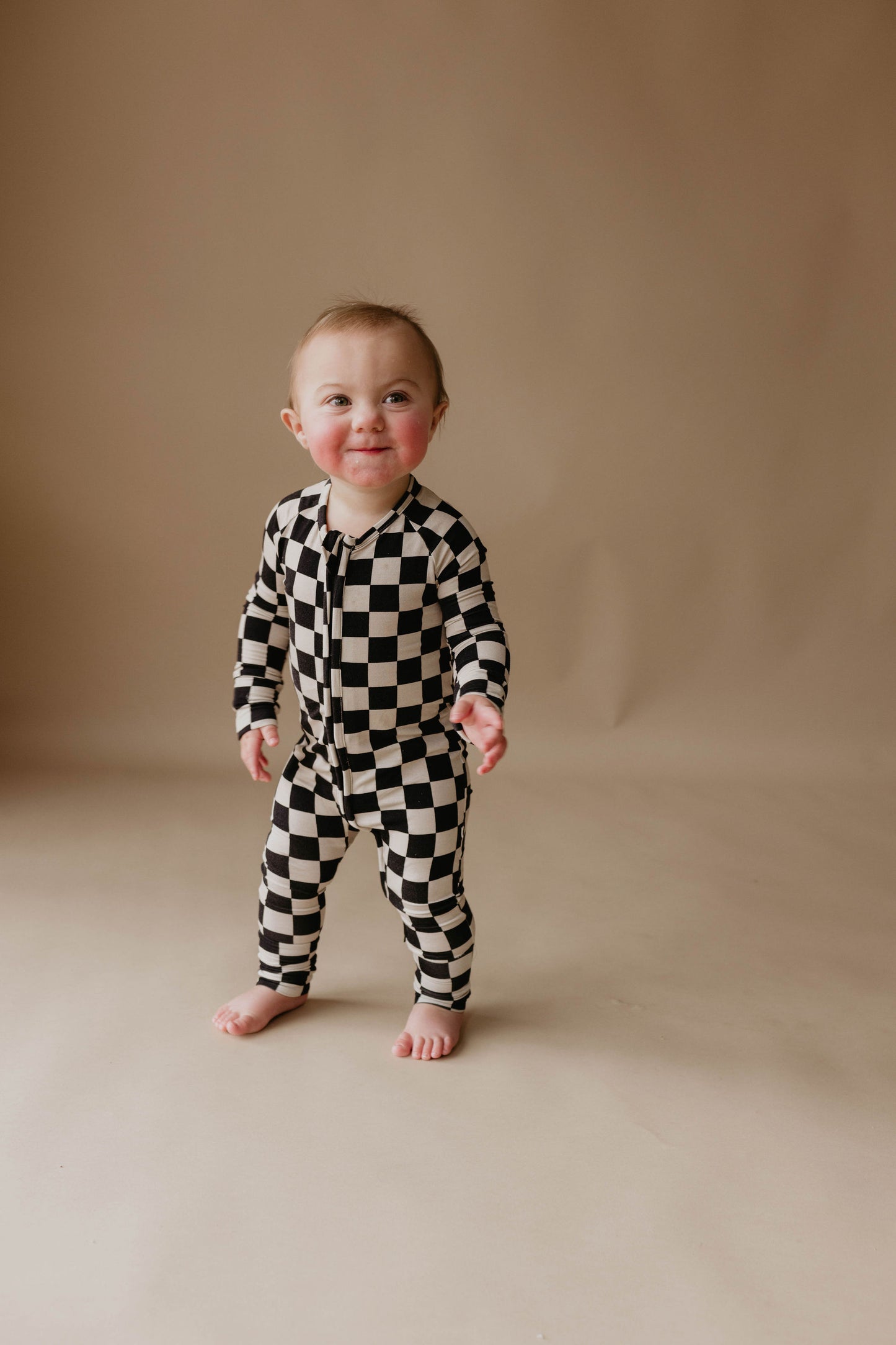 A smiling baby stands on a neutral-colored floor wearing the Bamboo Zip Pajamas in Black Checkerboard from forever french baby. The plain beige background highlights the baby's cheerful expression and the convenient rollover cuffs of their premium bamboo pajamas.