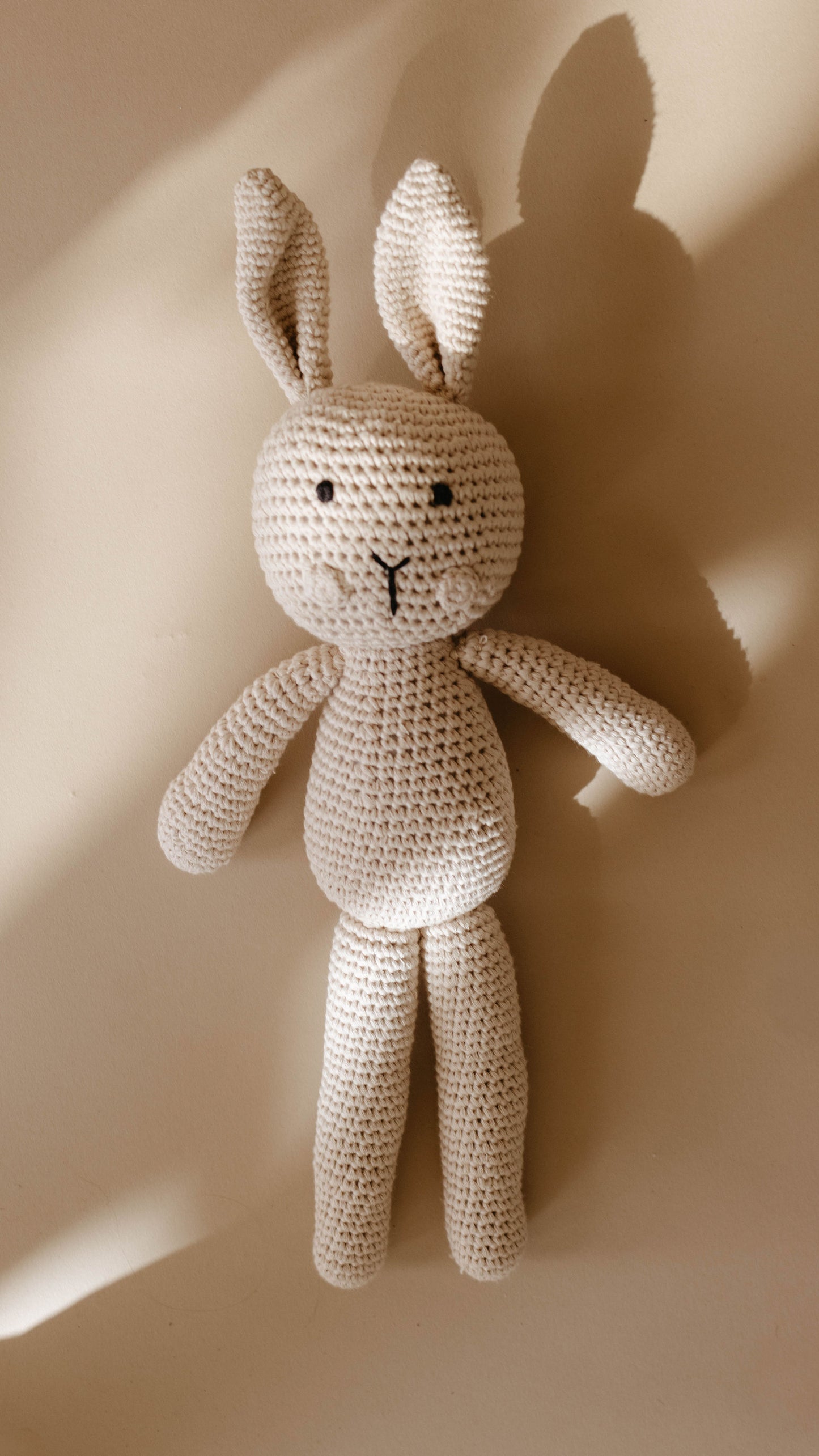 Honey the Bunny, a hand-crocheted knit doll from forever french baby, lies on a light beige surface. Crafted with light beige yarn, this charming stuffed bunny features long ears, small black eyes, and a stitched mouth. It casts a soft shadow and exudes the timeless charm of a Forever French Baby treasure.
