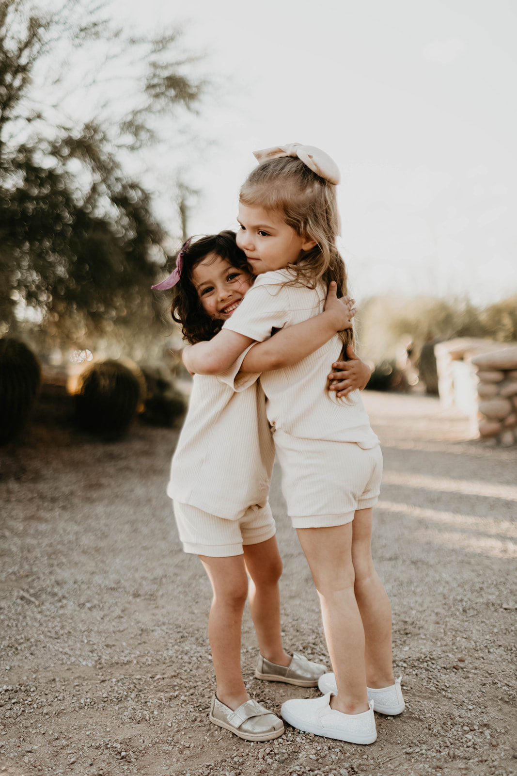 Two young girls are on a gravel path in a warm outdoor setting. Both dressed lightly, one in the Ribbed Set - Vanilla by forever french baby, they embrace while one smiles brightly. Trees and a stone structure blur softly in the background.