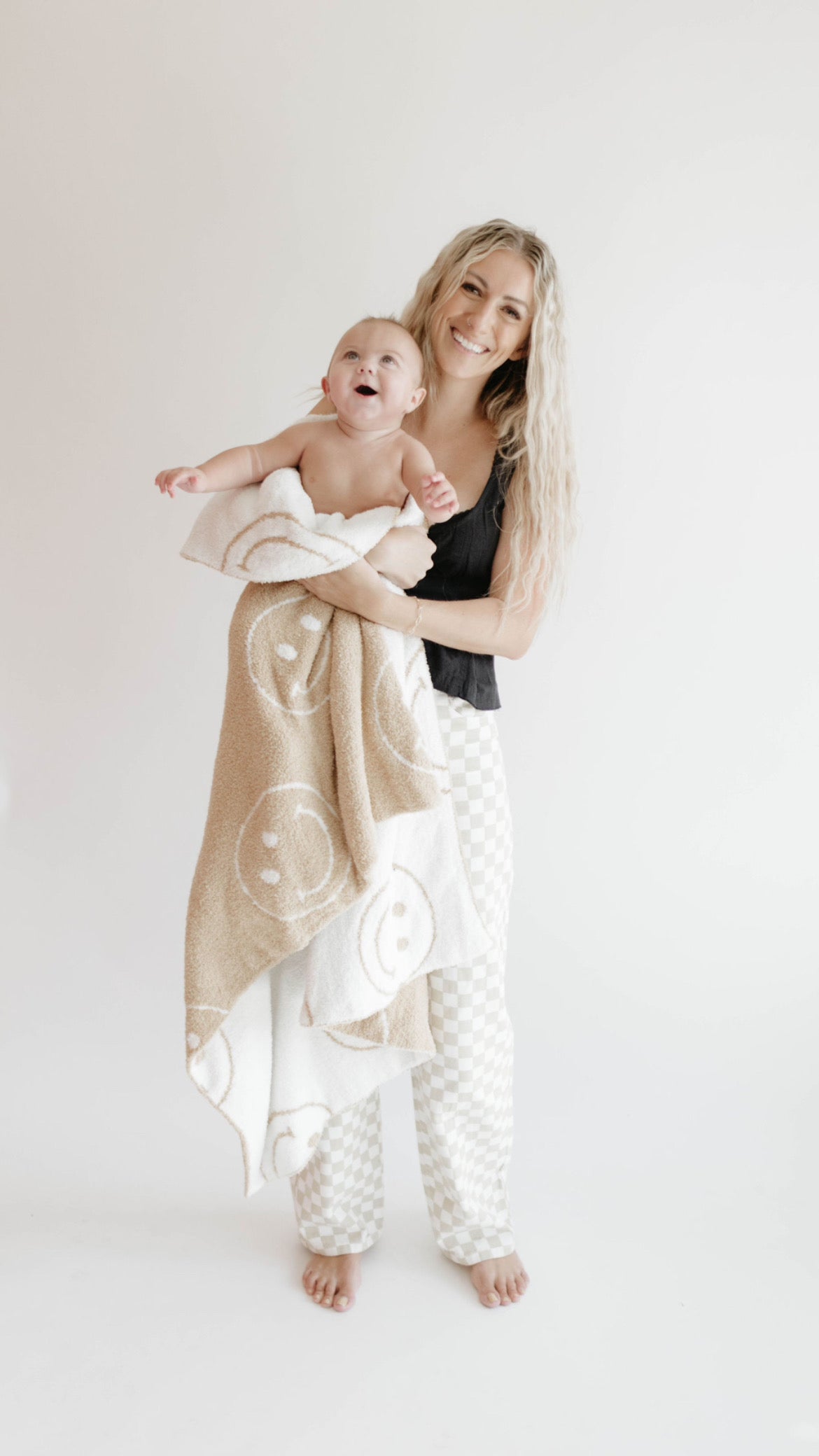 A smiling woman with long blonde hair holds a happy baby wrapped in the forever french baby Plush Blanket | Original Just Smile, featuring dreamy soft Oeko-tex certified fabric with white smiley faces. They stand barefoot against a plain white background.