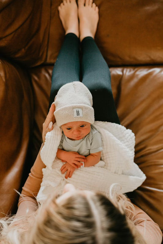 A person with blonde hair, viewed from above, cradles a newborn baby wrapped in a white blanket. The baby wears an ff Knit Beanie in Oatmeal by forever french baby and a light blue onesie while lying on the person's lap on a brown leather couch.