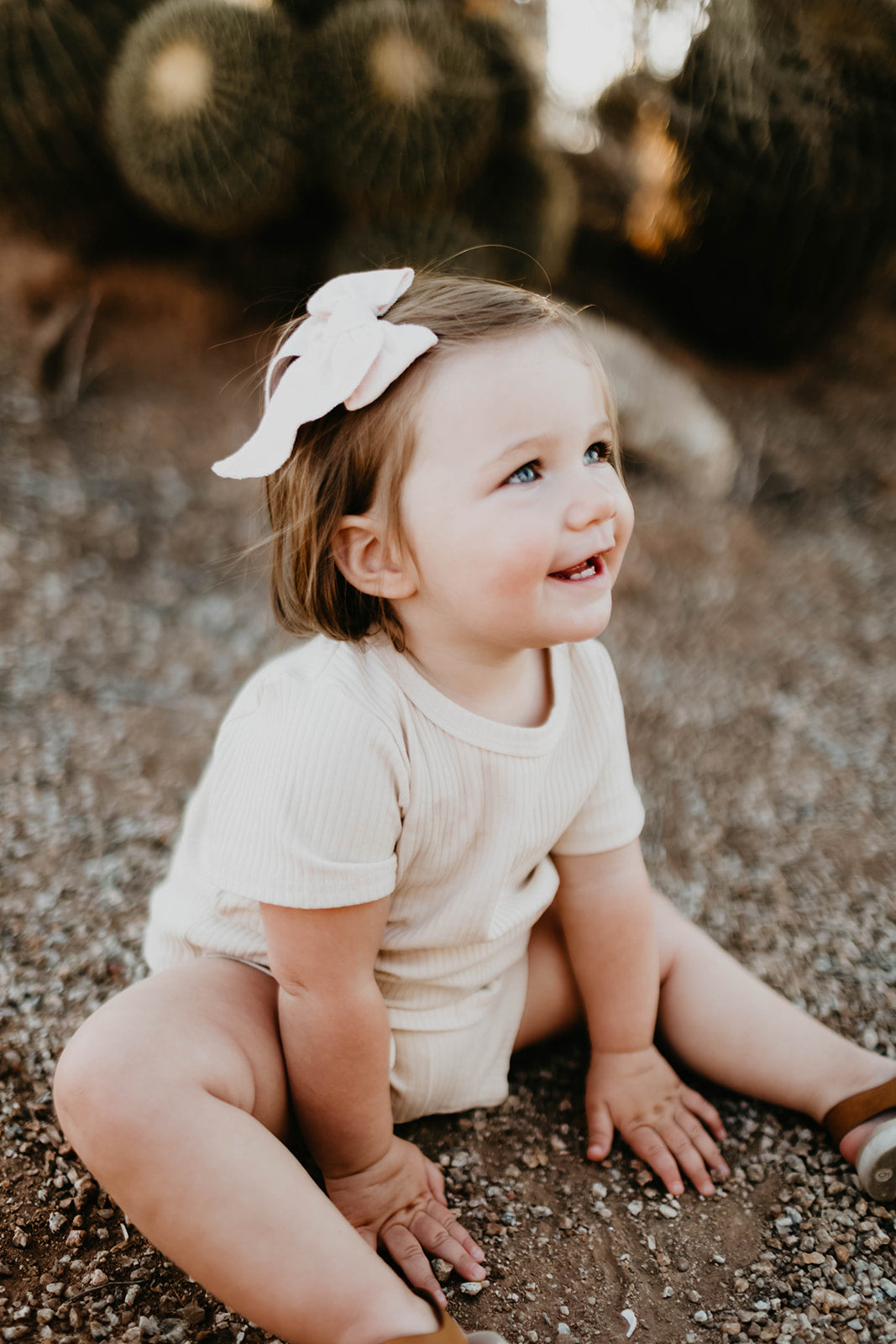 A toddler with short brown hair and a pale pink bow sits on gravel, wearing the Ribbed Set - Vanilla by forever french baby. The child is smiling and looking up, surrounded by cacti in the background.