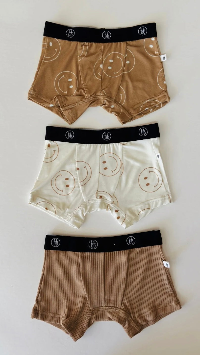 Three pairs of Bamboo Boxers from forever french baby are arranged vertically on a flat surface. The top pair is tan with smiley faces, the middle pair is ivory with tan smiley faces, and the bottom pair is solid beige with a ribbed texture. Each boxer features a black waistband adorned with the circular logo from Forever French Baby and ensures hypo-allergenic comfort.