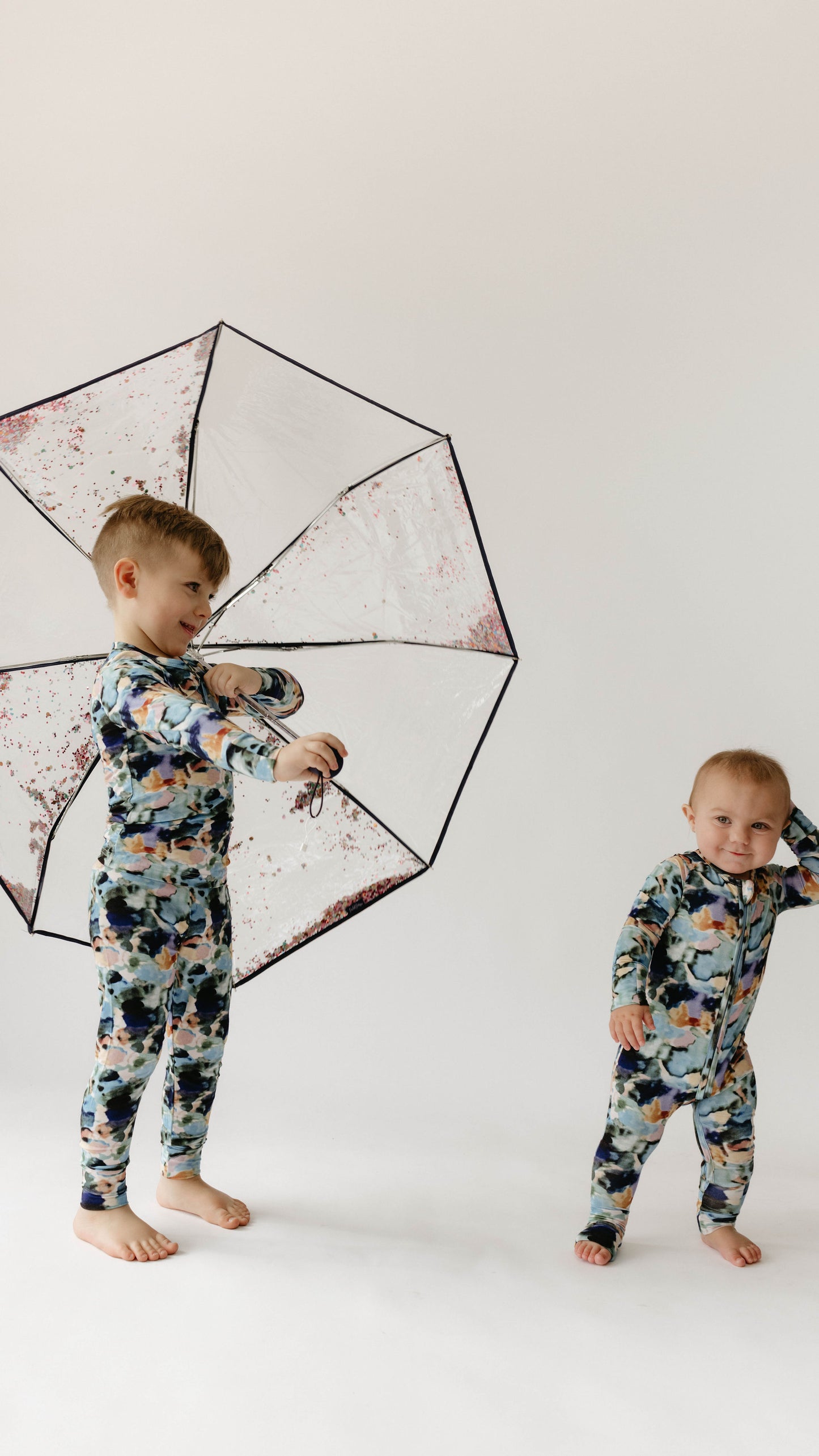 Two young children, both wearing matching "Bamboo Zip Pajamas | Charli Print" from forever french baby, stand against a plain white background. The older child holds a transparent umbrella with colorful accents, while the younger child stands close, smiling and holding their head with one hand.