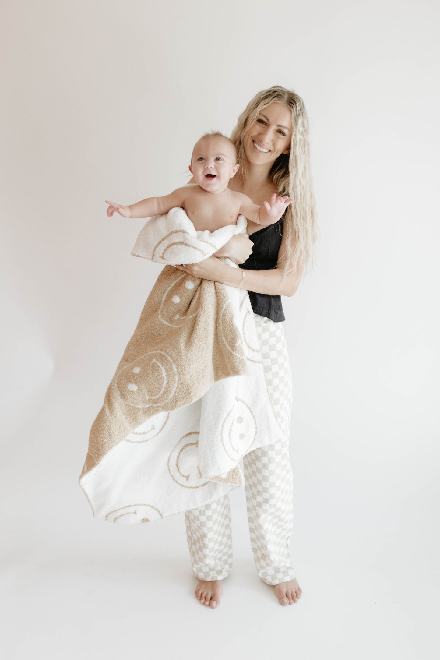 A smiling woman with long, wavy blonde hair holds a happy baby wrapped in the forever french baby Plush Blanket | Original Just Smile, made from Oeko-Tex certified Microfiber feather yarn. Both are barefoot against a plain white background.
