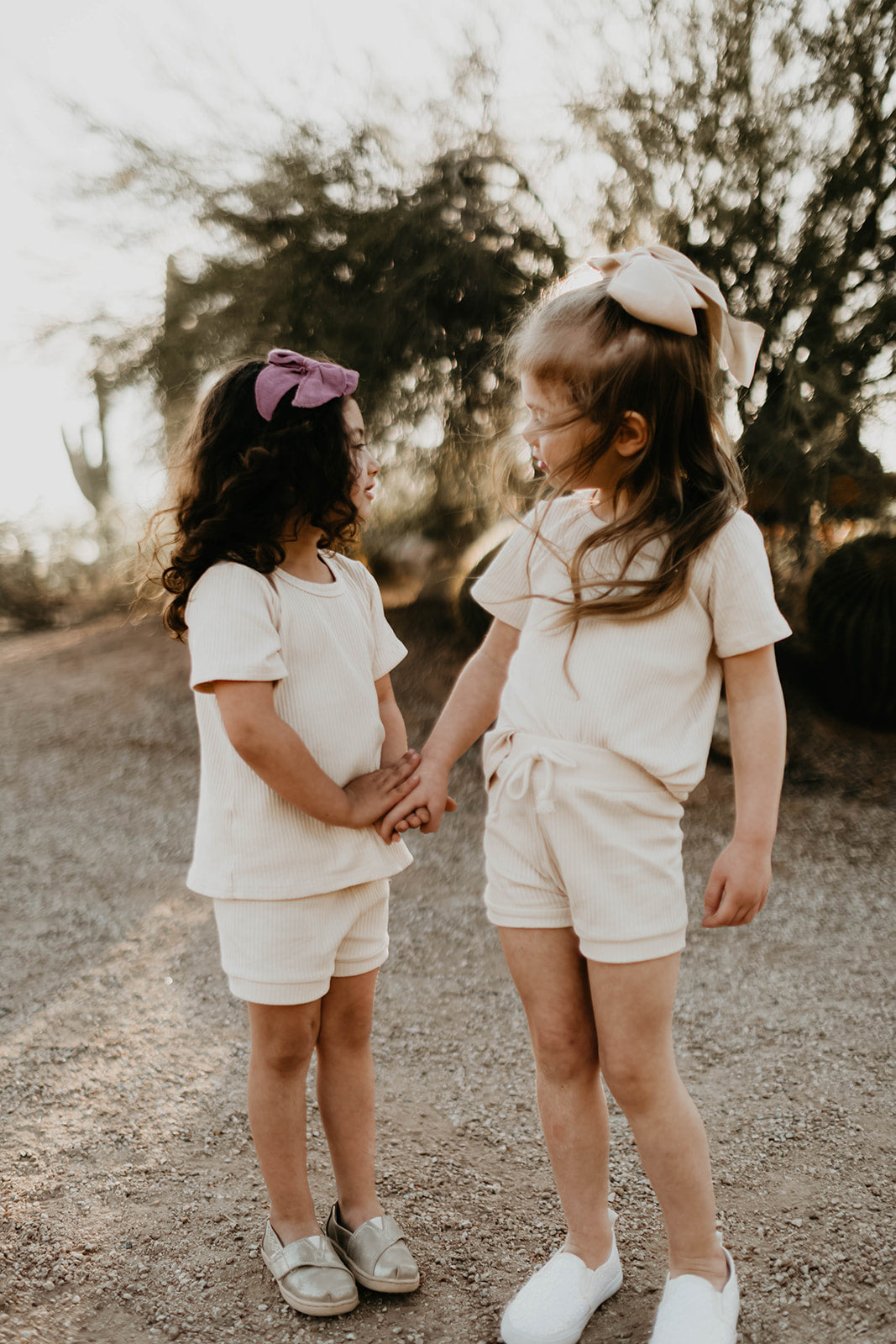 Two young children stand outdoors in matching "Ribbed Set - Vanilla" by forever french baby, holding hands; one wears a purple bow, the other pink. Lush trees frame their tender moment.