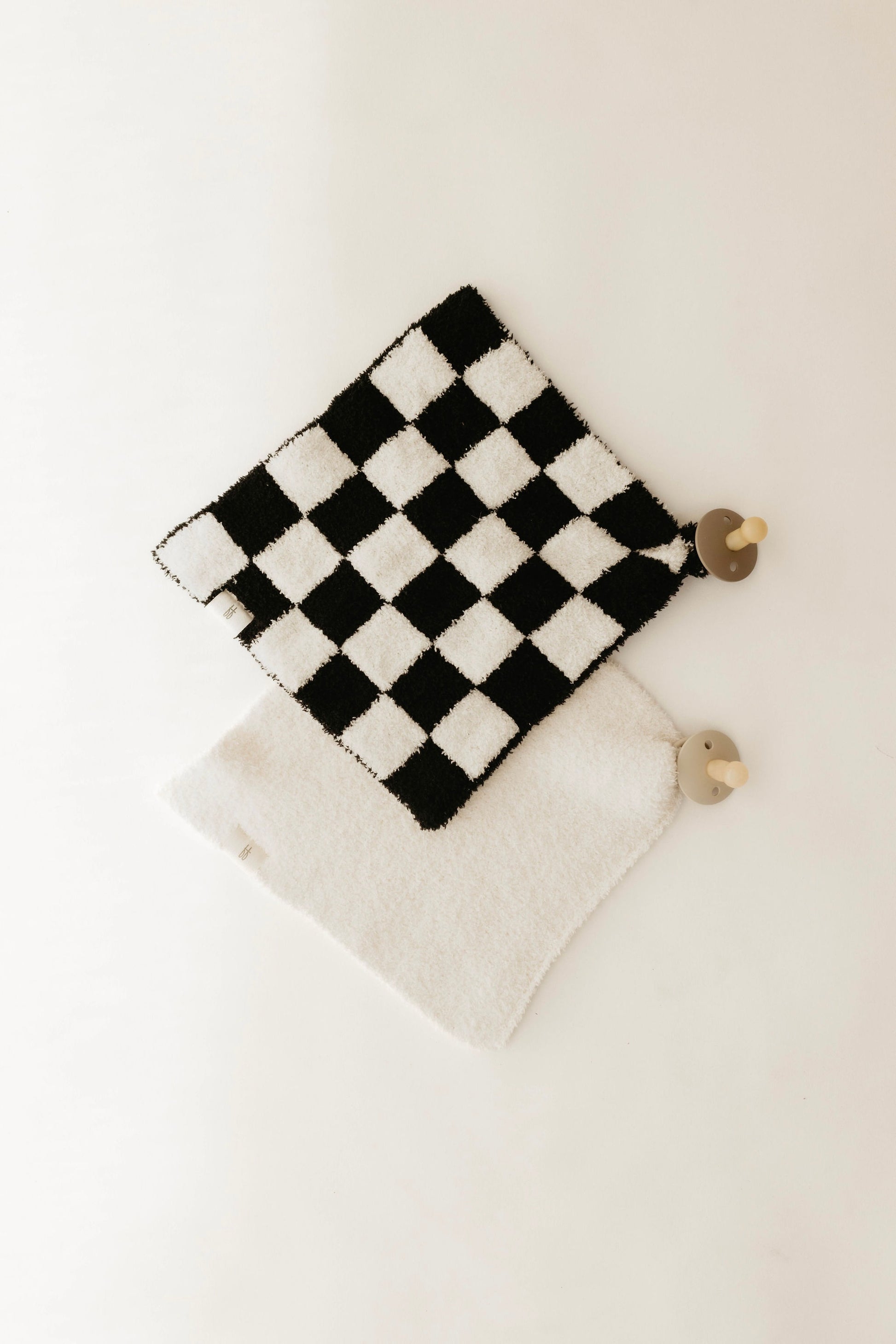 A Lovey by forever french baby, featuring a black and white checkerboard pattern, and a plain white potholder are laid on a light surface. Each is crafted with microfiber feather yarn and features a wooden hook at the corner for hanging.
