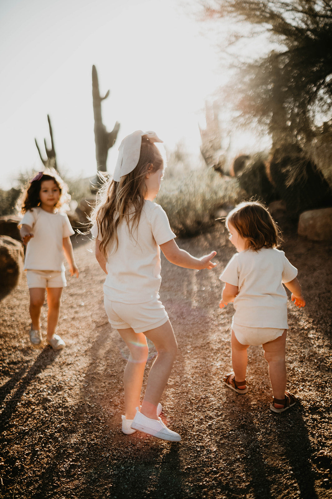 Three children in oversized, light-colored Ribbed Set - Vanilla by forever french baby walk and play on a sunlit desert path, with cacti and trees in the background under a warm sunlit glow.