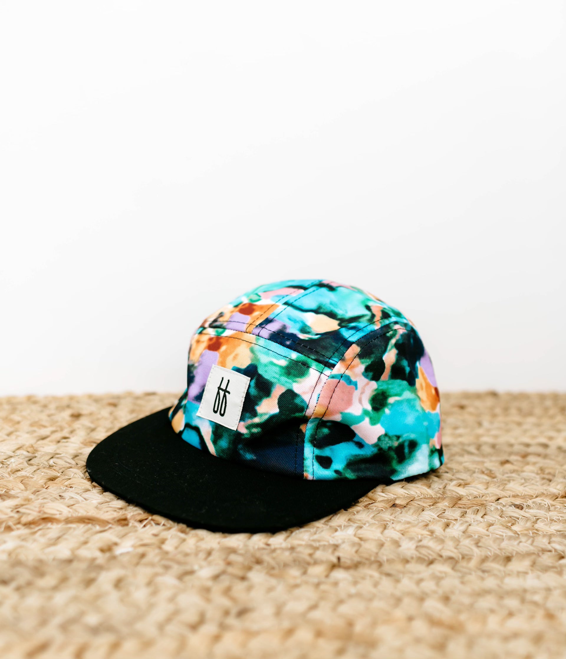 The Snap Back | Charli Print by forever french baby features an exclusive and vibrant design with blue, green, orange, and purple tones paired with a black bill. Placed on a woven surface, this colorful baseball cap showcases a small white logo with black markings on the front. Part of a limited collection, it stands out beautifully against the plain white background.