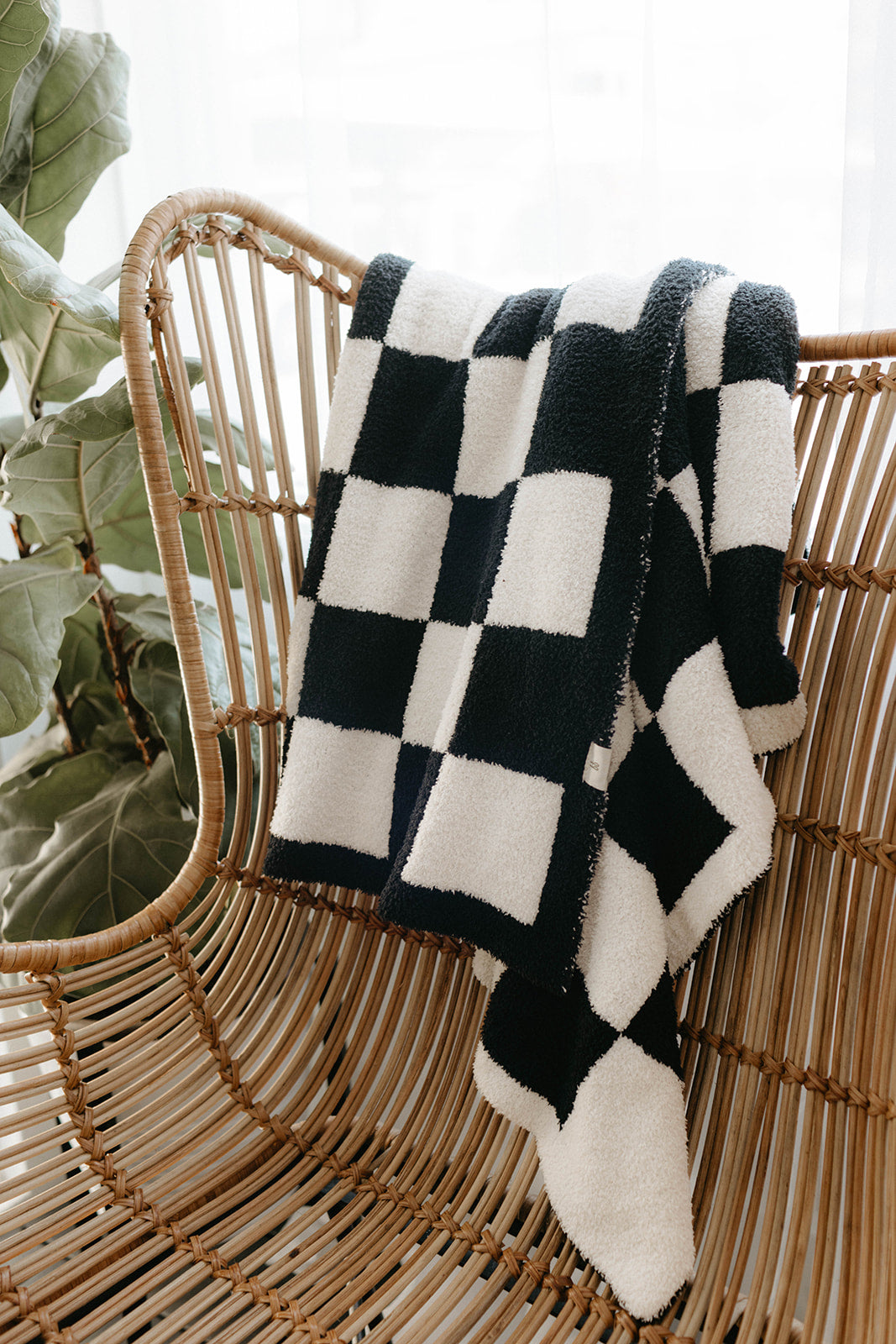 A black and white checkerboard "Plush Blanket" from forever french baby, OEKO Standard 100 certified for safety, is casually draped over a woven rattan chair. The chair is positioned near a window with sheer curtains, allowing soft natural light to illuminate the scene. A large leafy plant is partially visible on the side.