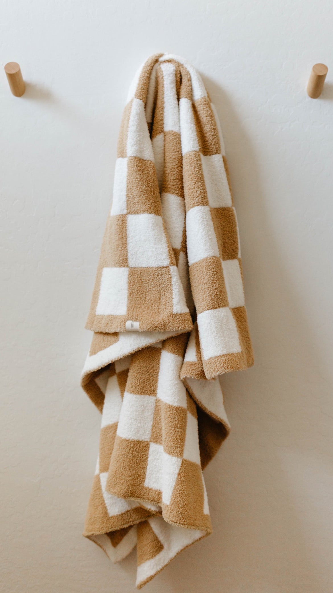 A plush beige and white checkered towel from the "Original Checkerboard" collection by Forever French Baby hangs on a wooden peg against a light-colored wall. Two pegs are visible; the towel occupies one while the other remains empty. The soft texture, crafted from microfiber feather yarn, and the neat checkerboard pattern create a cozy and inviting appearance.