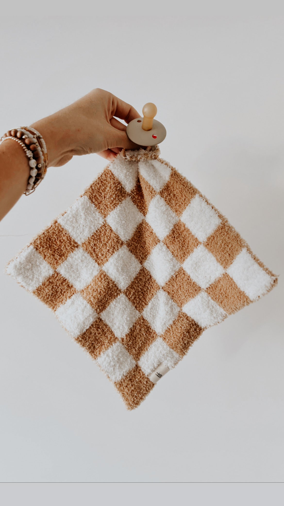 A hand adorned with a beaded bracelet holds a wooden peg, from which hangs the Lovey | Original Checkerboard from forever french baby, featuring soft white and light brown checkered squares made of luxurious microfiber feather yarn against a plain background.