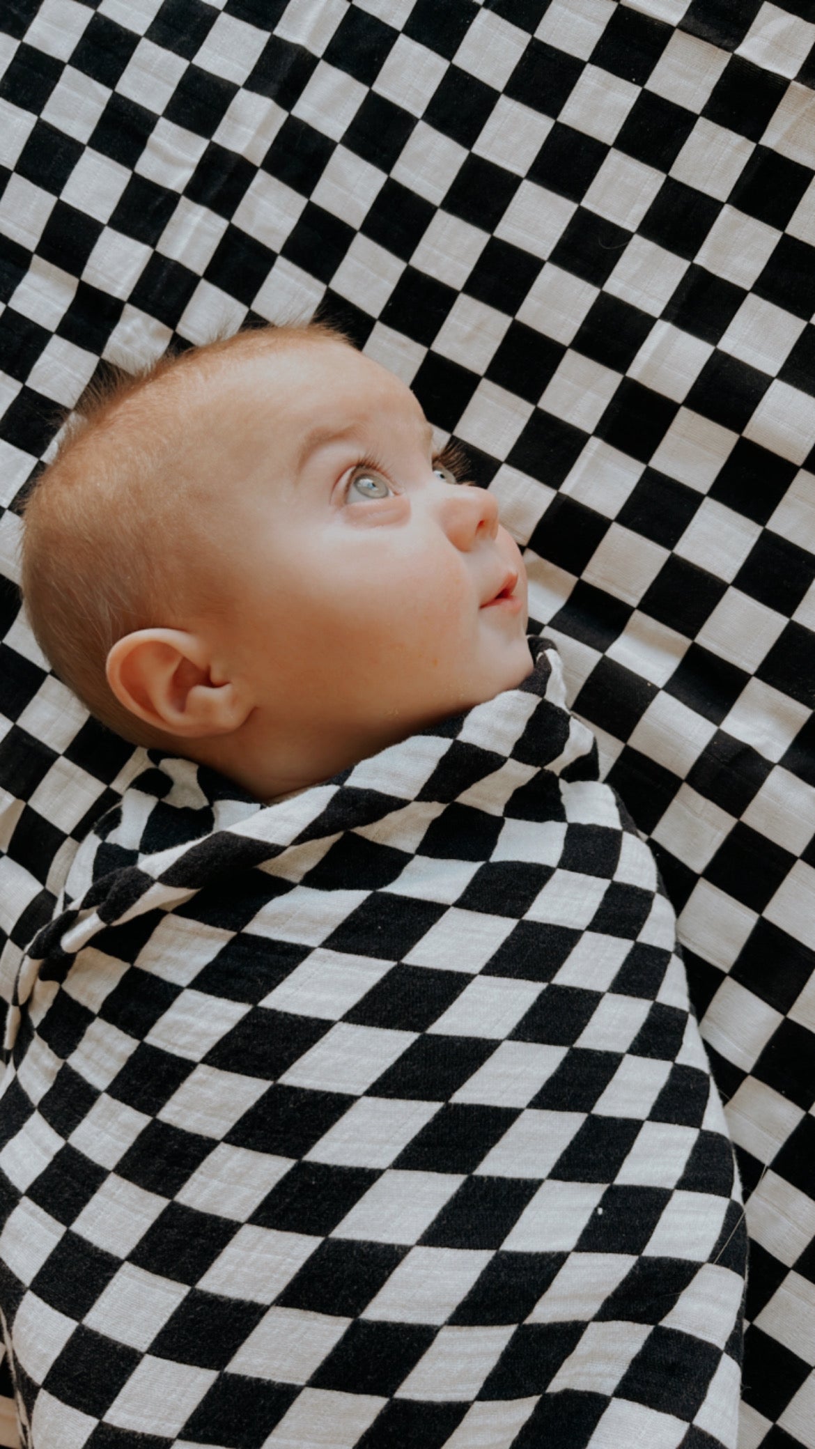 A baby wrapped in a forever french baby Muslin Swaddle | Black & White Checkerboard lies on a matching checkered surface, gazing upward with wide eyes.