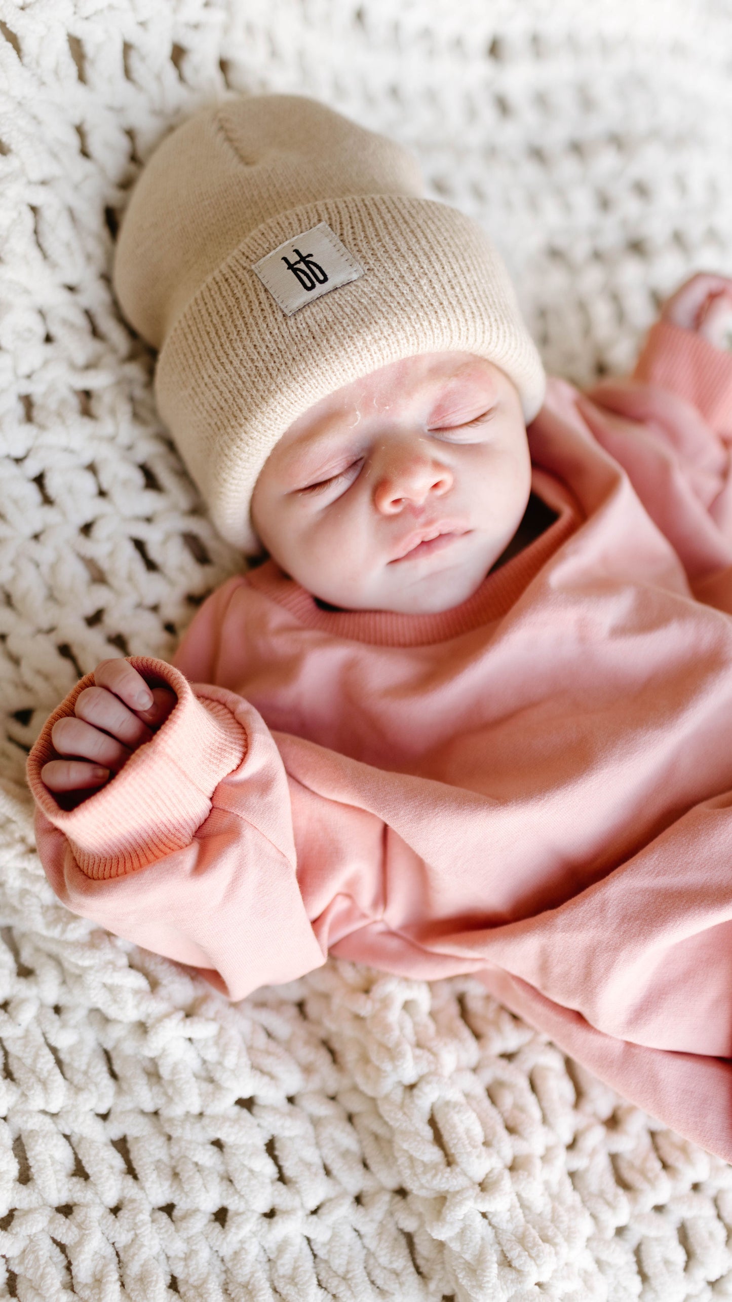 A newborn wearing the forever french baby Sweatshirt Romper in Rose and a beige knit hat lies on a chunky white knitted blanket, eyes closed and hands near the face.