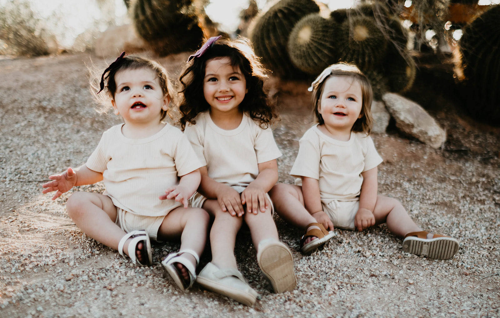 Three young children sit on gravel in a desert setting, smiling and wearing forever french baby's Ribbed Set - Vanilla with sandals. Cacti are visible in the background as the children appear happy and engaged, dressed in light-colored clothing.
