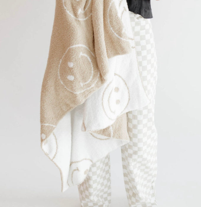 A person in light-colored checkered pants holds a beige and white forever french baby Plush Blanket | Original Just Smile, featuring large smiley faces. The background is plain white.