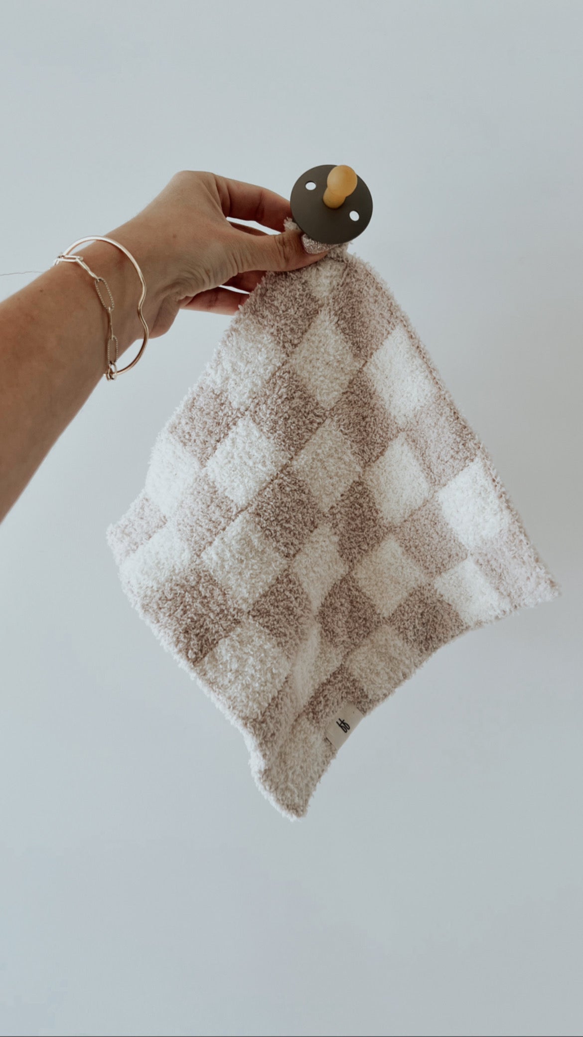 A person's hand holding the Lovey in Taupe & White Checkerboard by forever french baby, featuring an attached soft blanket with a neutral checkerboard pattern. The blanket showcases Forever French Baby's microfiber feather yarn for ultimate softness. The hand also wears a delicate bracelet, set against a plain white background.