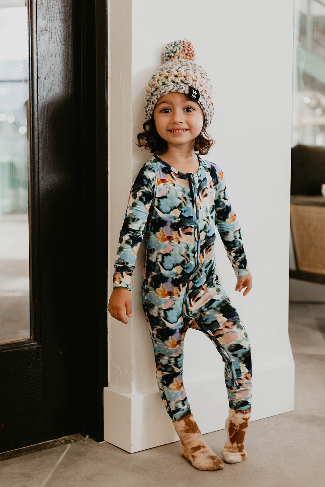 A young child with curly hair stands leaning against a wall, smiling. They are wearing vibrant Bamboo Zip Pajamas in the Charli Print from forever french baby, made from soft bamboo fabric, along with a thick knitted beanie crafted from multicolored yarn.