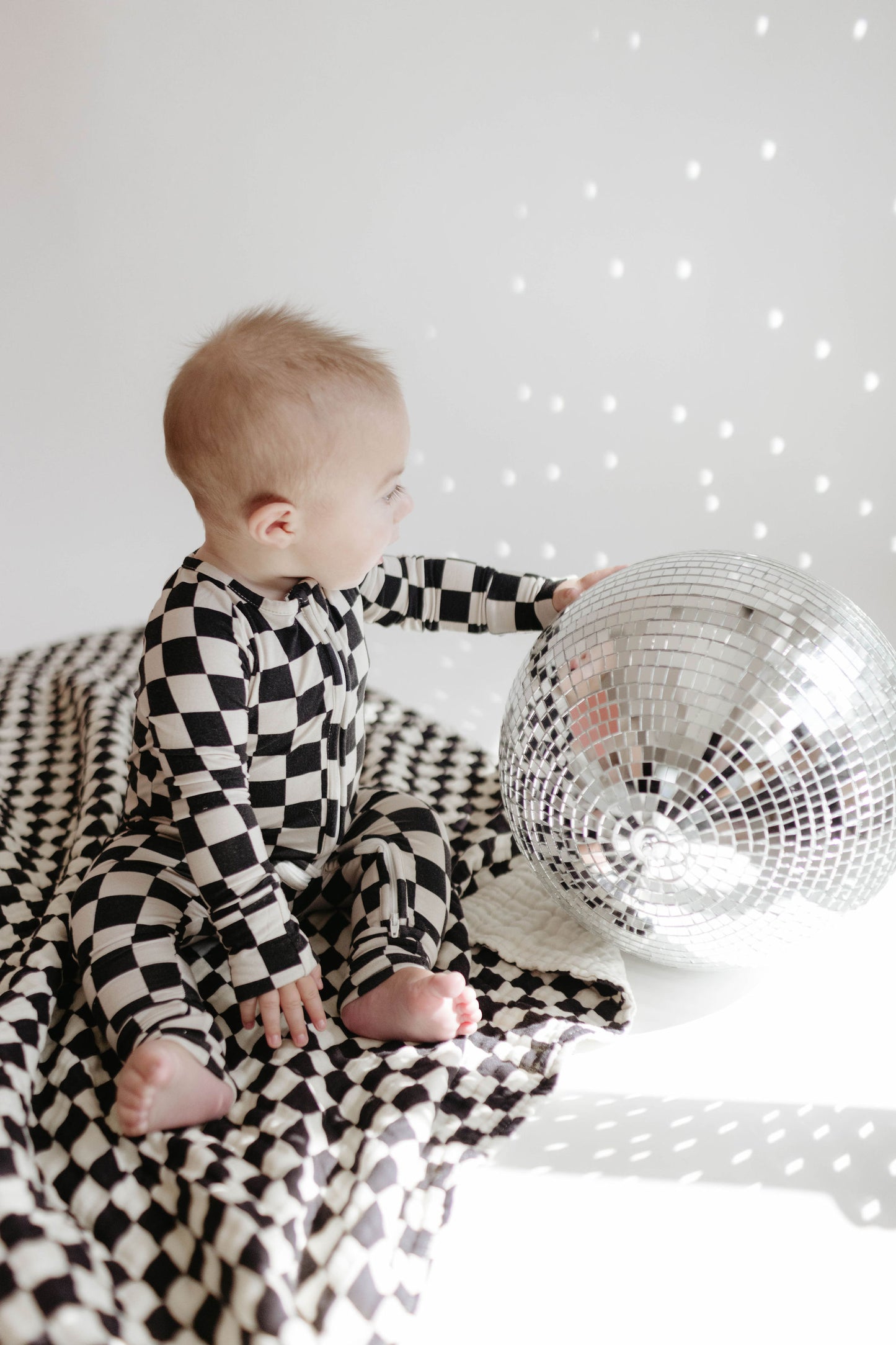 A baby wearing a charming outfit sits on the *Muslin Quilt in Black & White Checkerboard* from **Forever French Baby**, reaching out to touch a shiny disco ball. The background is white, and the disco ball casts small reflective light spots around the scene.
