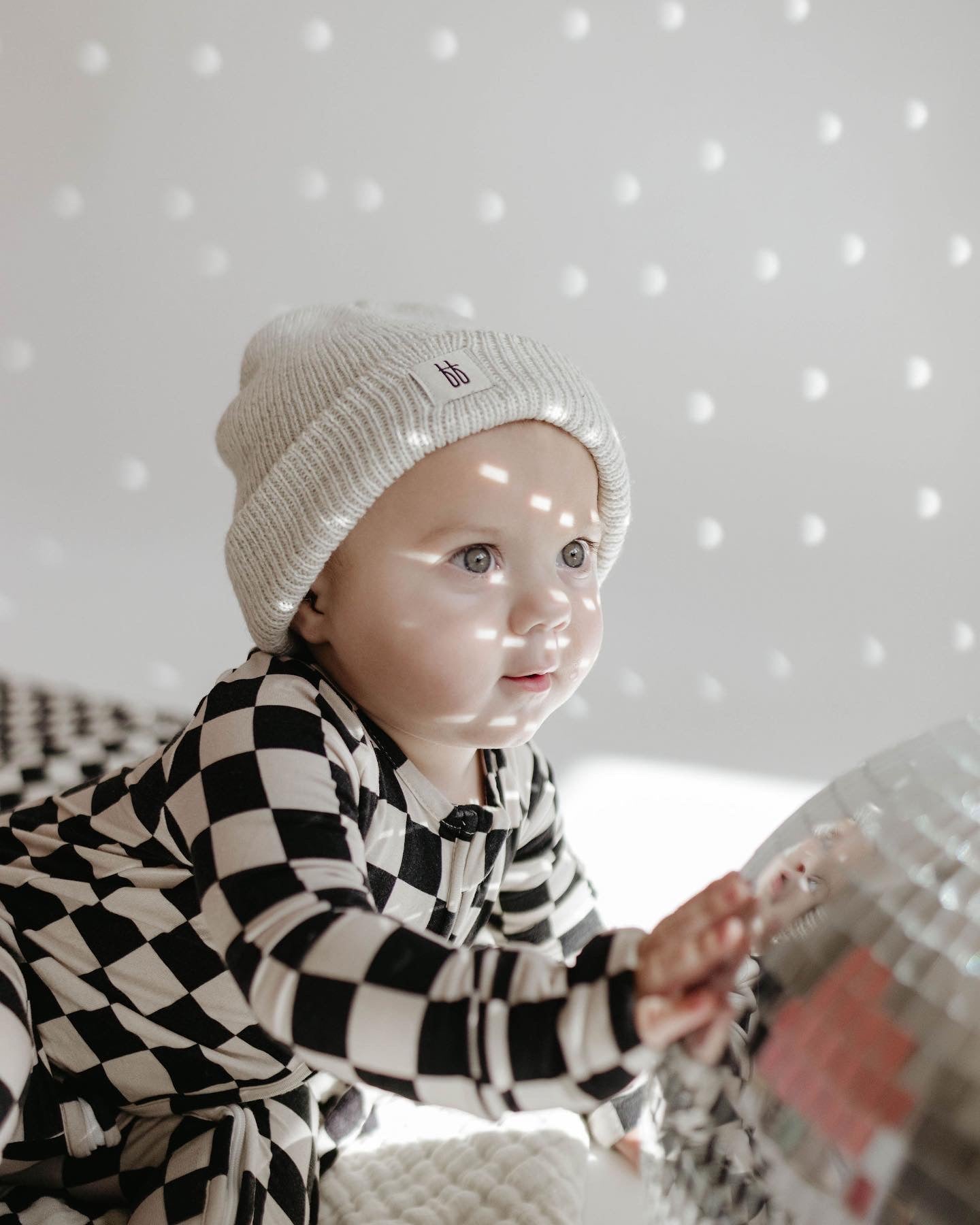 A baby wearing Forever French Baby's Bamboo Zip Pajamas in a black checkerboard pattern and a light gray knit hat sits on a patterned surface, touching a large mirrored disco ball. Soft lighting creates a gentle and cozy atmosphere with small light reflections on the baby's face and background.