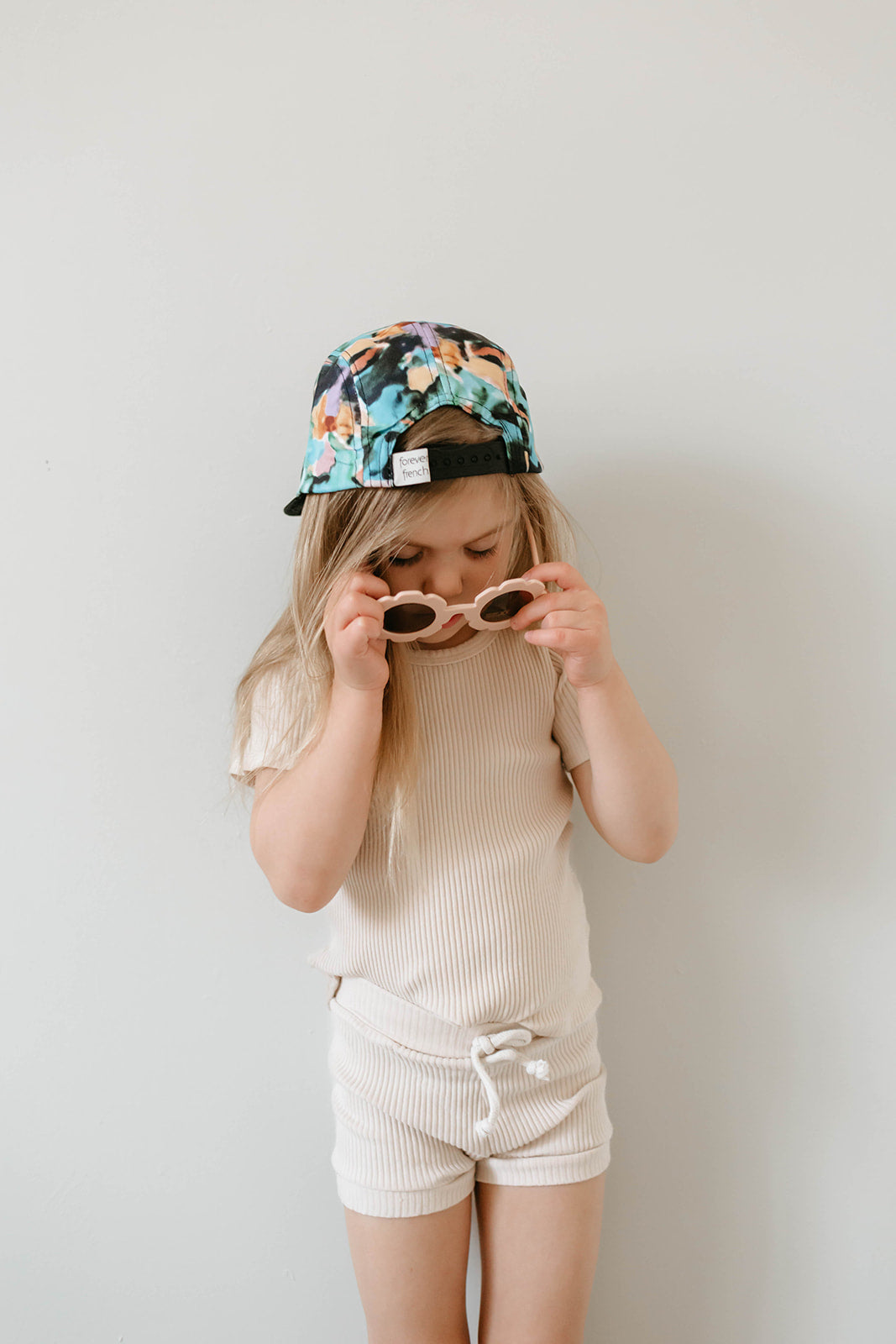 A young child with long hair wears a Snap Back | Charli Print cap from forever french baby backward and a cream-colored ribbed outfit. The child, part of our limited collection, is looking down, adjusting a pair of beige sunglasses with both hands against a plain, light-colored background.