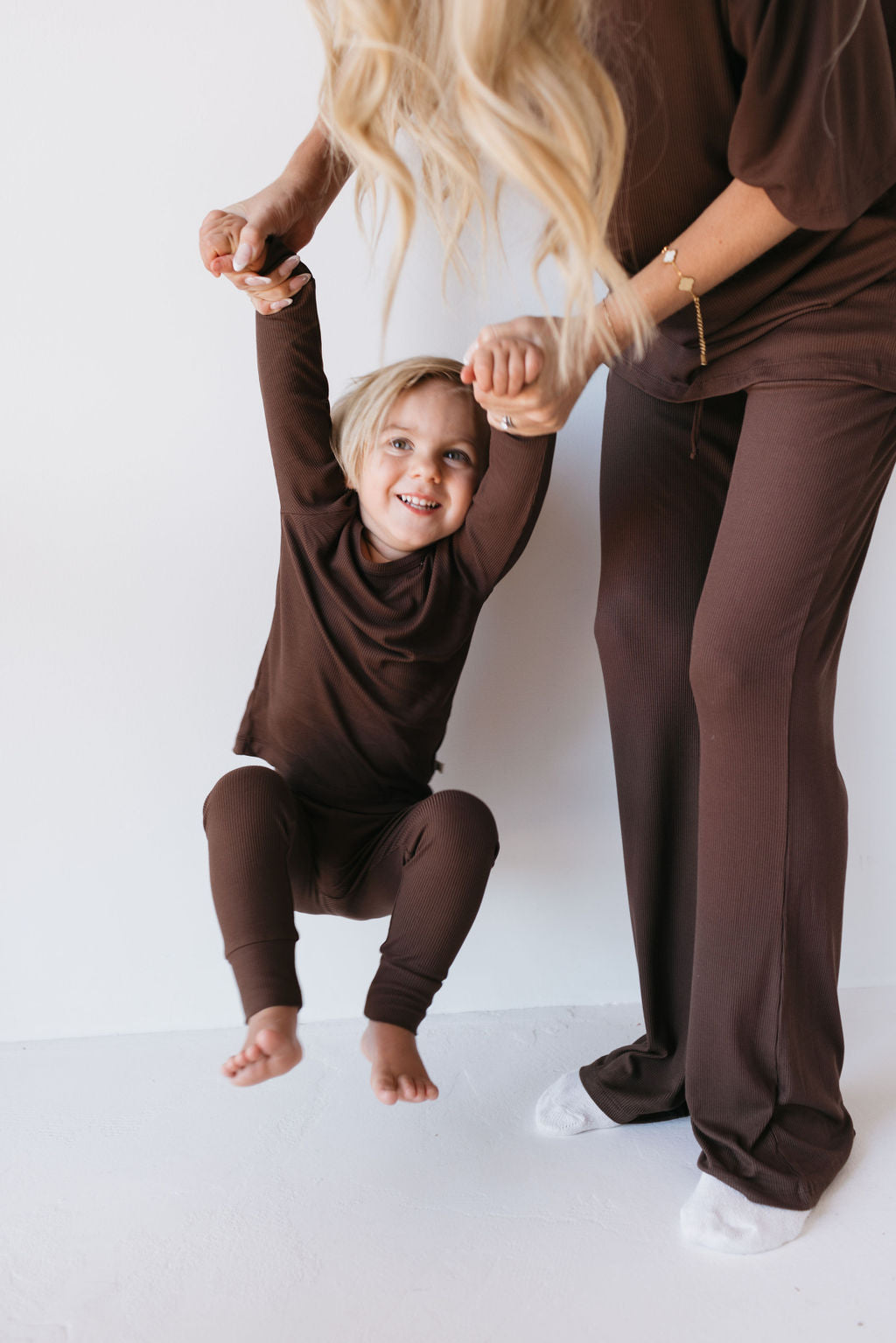 A child in brown pajamas playfully swings while holding a woman's hands. The woman, dressed in matching brown pants, stands on a white floor. The child smiles with excitement.