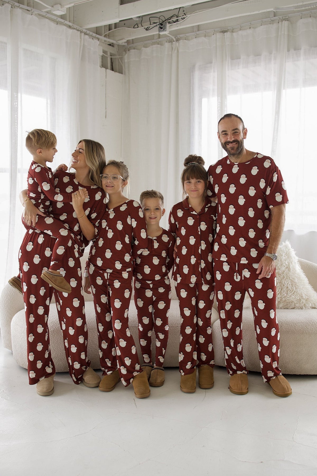 A family of six wearing matching maroon pajamas with a white snowman pattern stand together in a bright room.