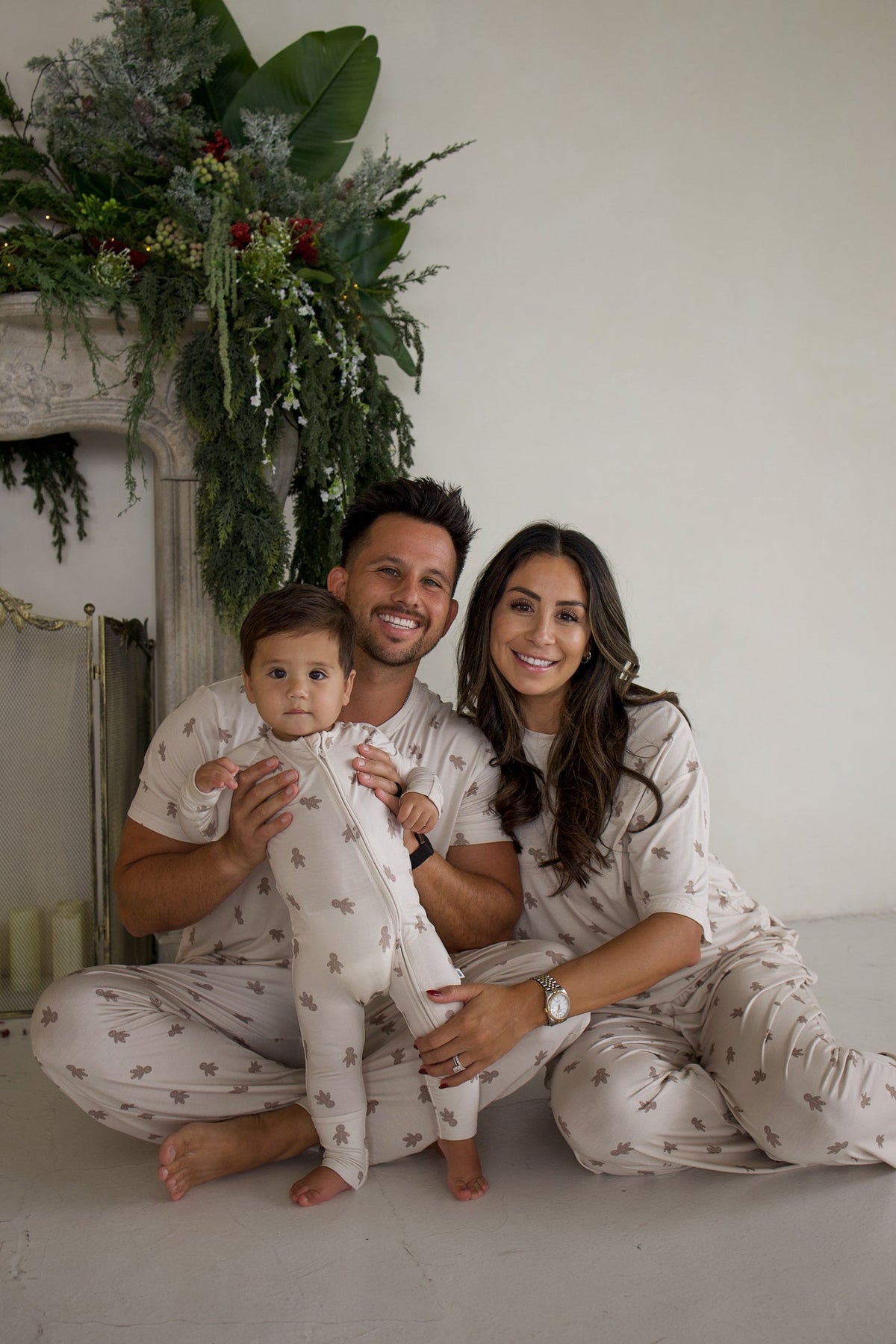 A family of three sitting together in matching beige gingerbread-patterned pajamas in front of a fireplace decorated with greenery.