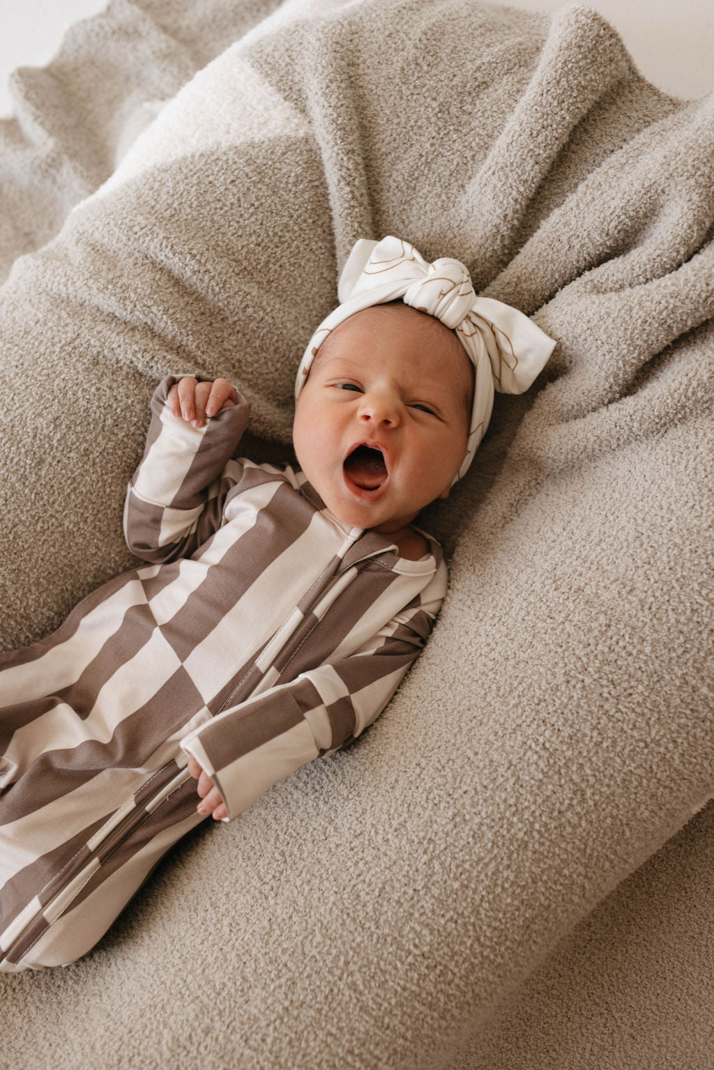 A baby wearing a brown and white striped onesie and a white headband is yawning while lying on a textured beige blanket.