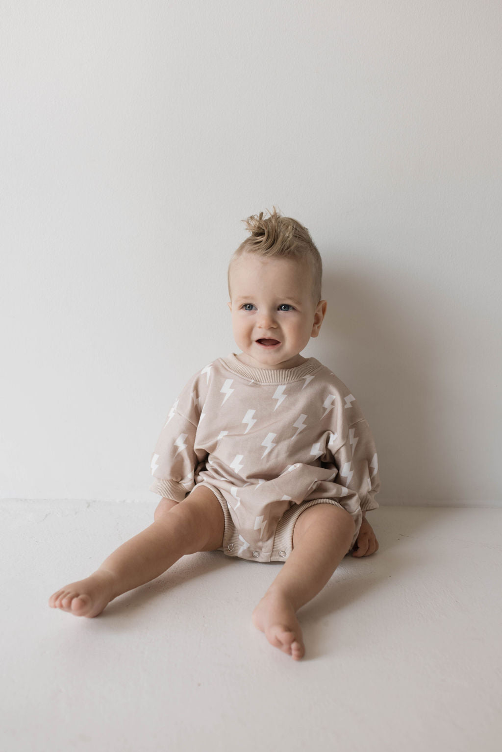 A baby with a light brown hairstyle sits on the floor against a white wall, wearing a beige outfit with white lightning bolt patterns. The child smiles and looks slightly to the side.