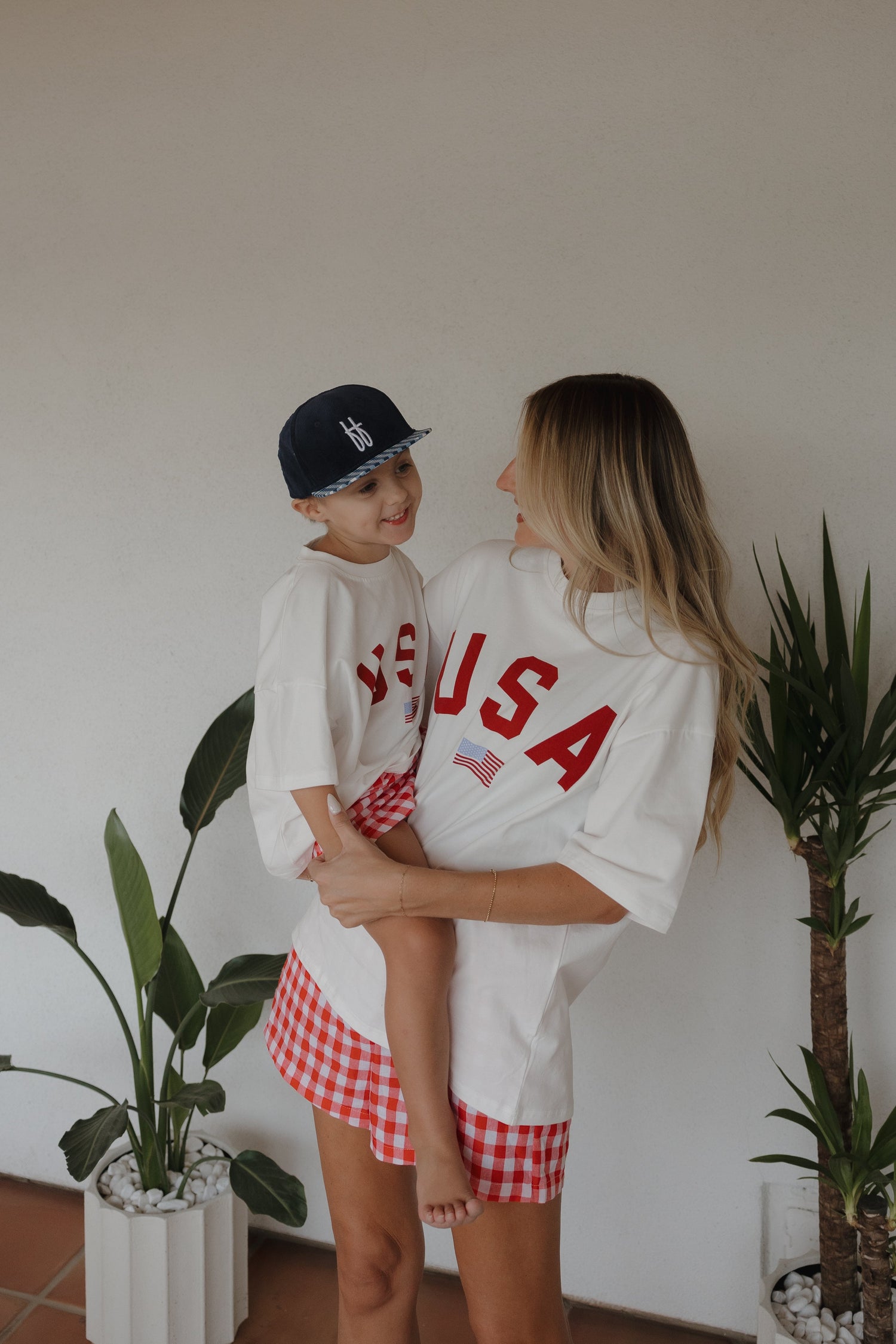 A smiling woman holds a young child, both wearing matching white USA t-shirts. The child also wears a navy hat. They stand indoors near green potted plants, and both have red checkered bottoms.
