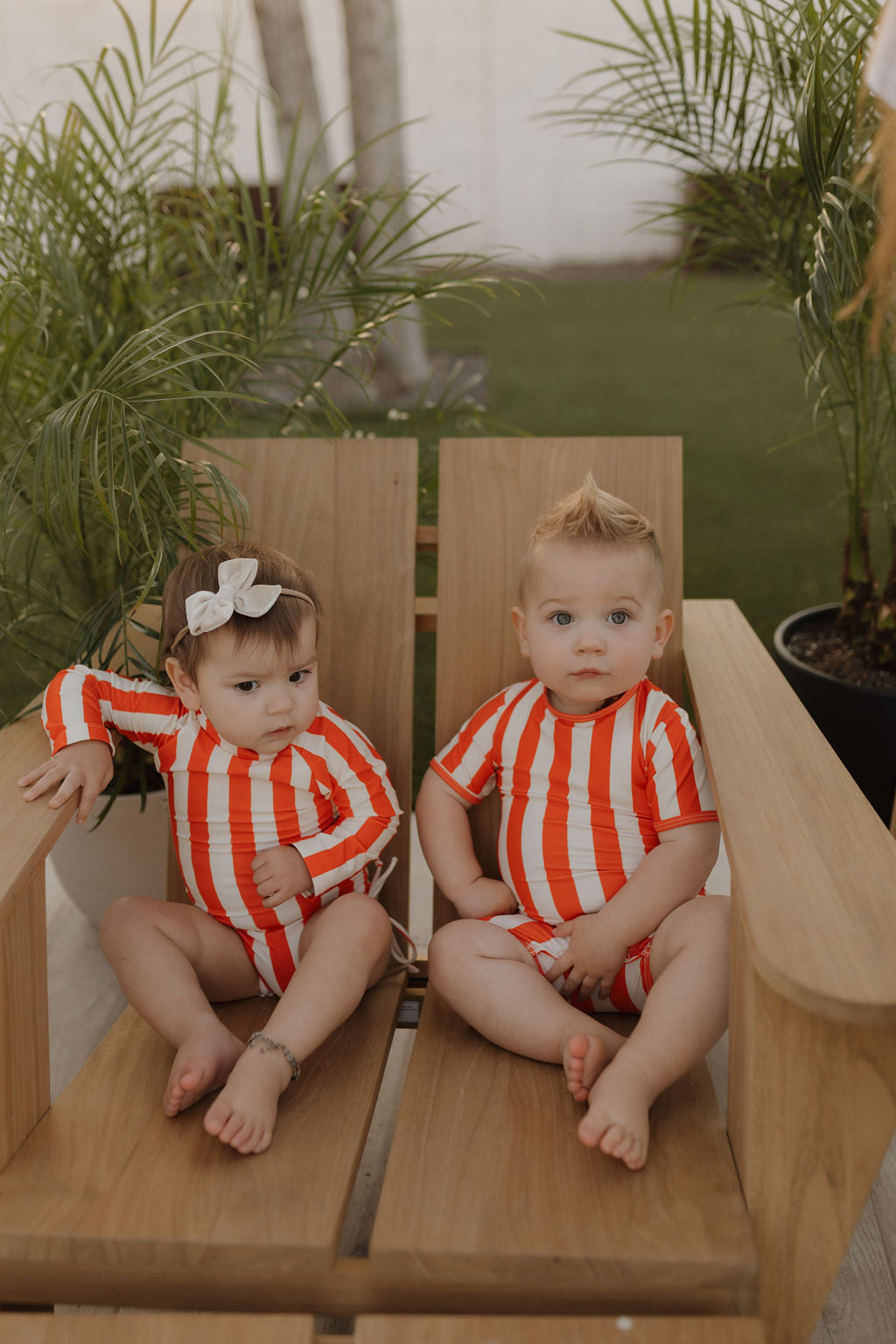 Two toddlers wearing matching red and white striped outfits sit on a wooden chair outdoors. One has a white bow in their hair. They are surrounded by potted plants on a sunny day.