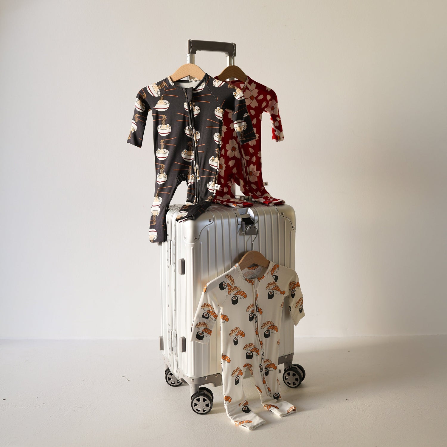 Three patterned baby onesies on hangers are displayed on and around a white suitcase with wheels against a plain, light background.