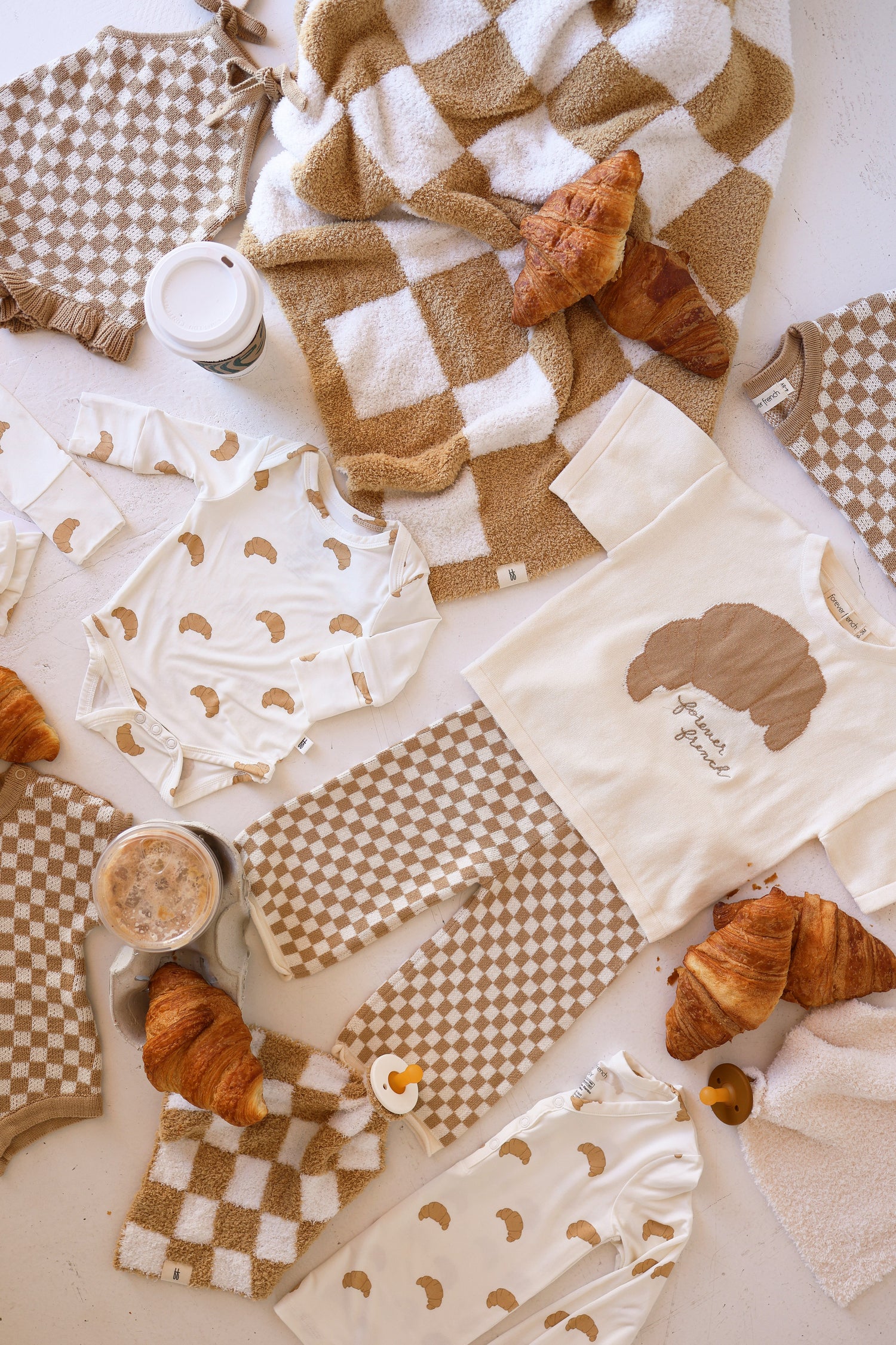 A flat lay features beige and cream baby clothes with checkered and croissant patterns, cozy blankets, coffee, and croissants scattered on a white surface.