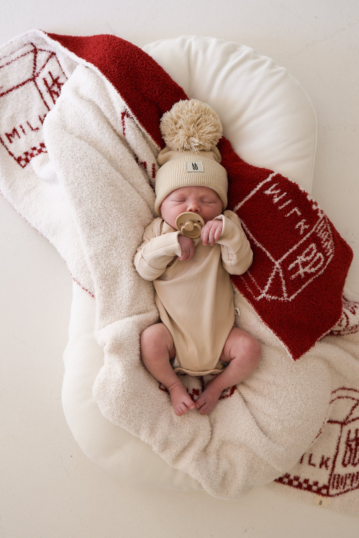 Newborn baby in a beige outfit and pompom hat lying on a soft cushion, swaddled with a cream and red blanket decorated with milk cartons, holding a pacifier.