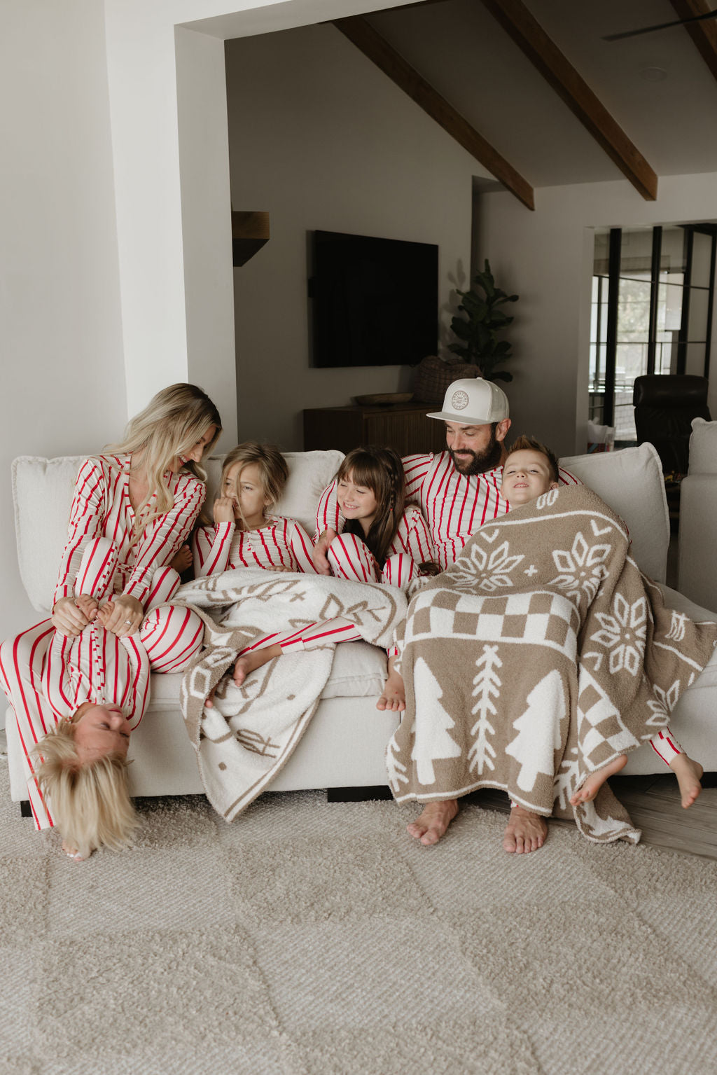 A family of six lounges on a sofa, wearing matching red and white striped pajamas. Some members are covered with a brown and white patterned blanket. One person is playfully hanging upside down.